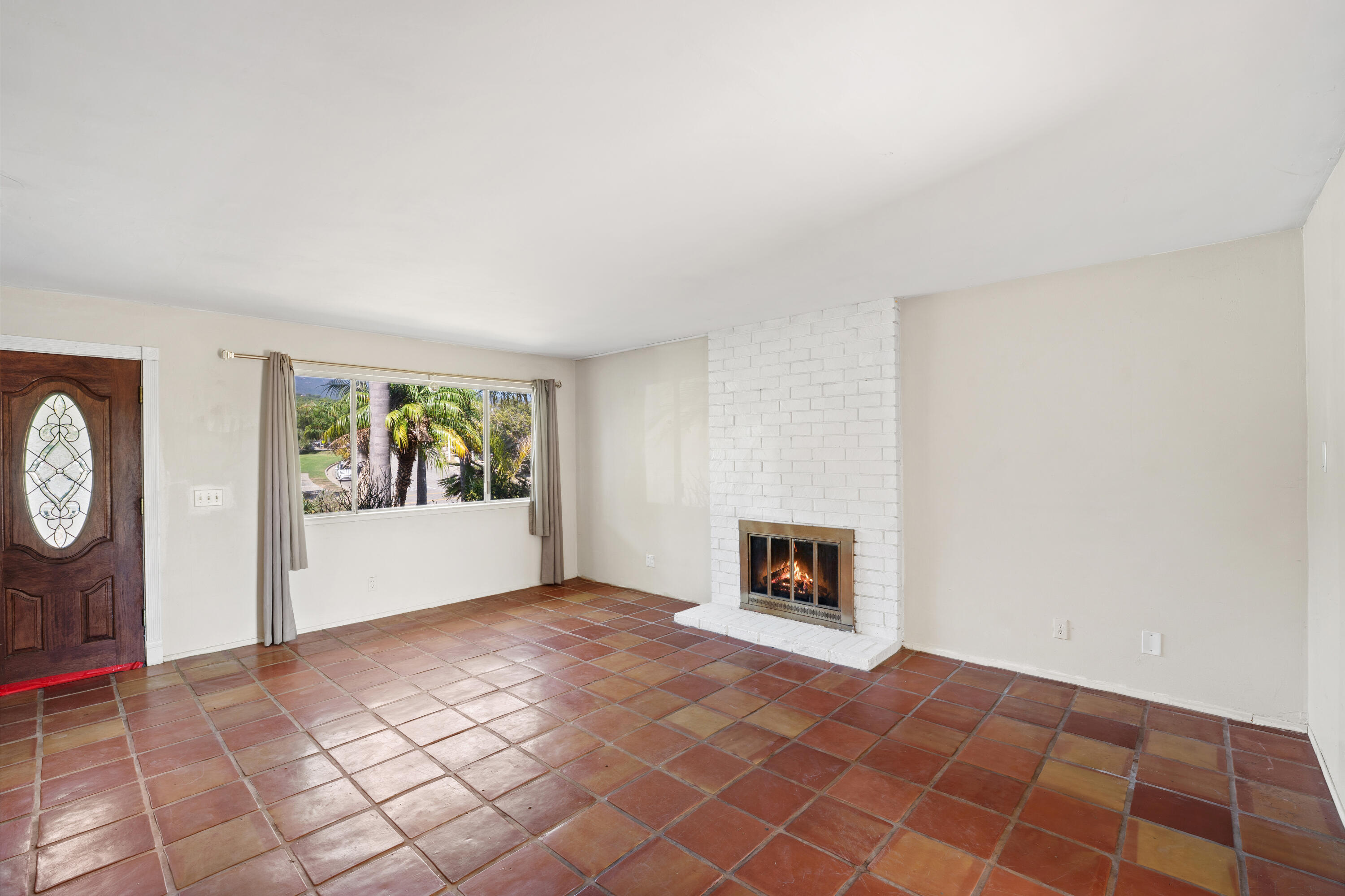 Unfurnished living room with terracotta tile floors, a lit white-brick fireplace, and a window overlooking palm trees.