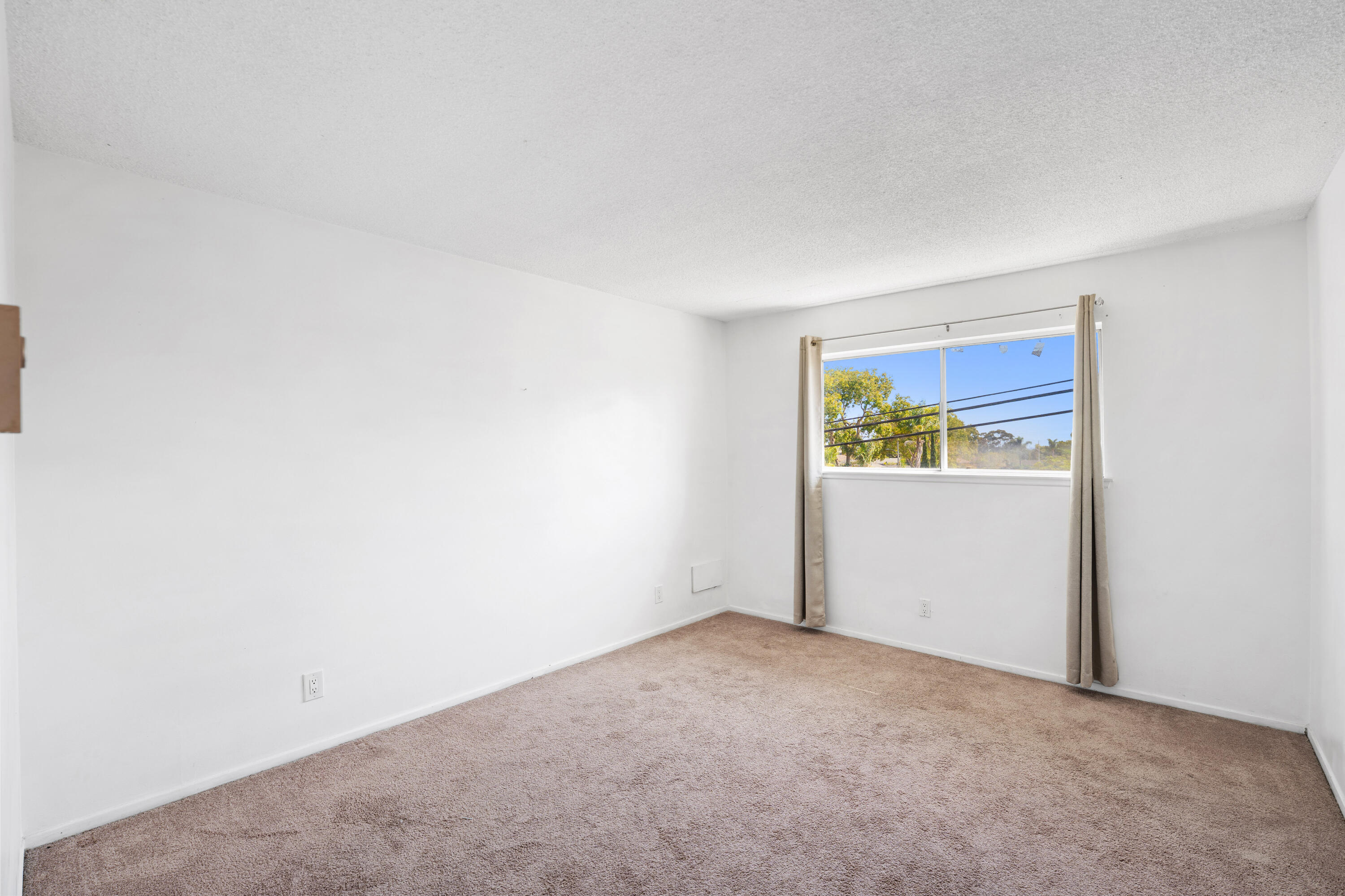 Empty carpeted room with white walls and a curtained window overlooking trees.
