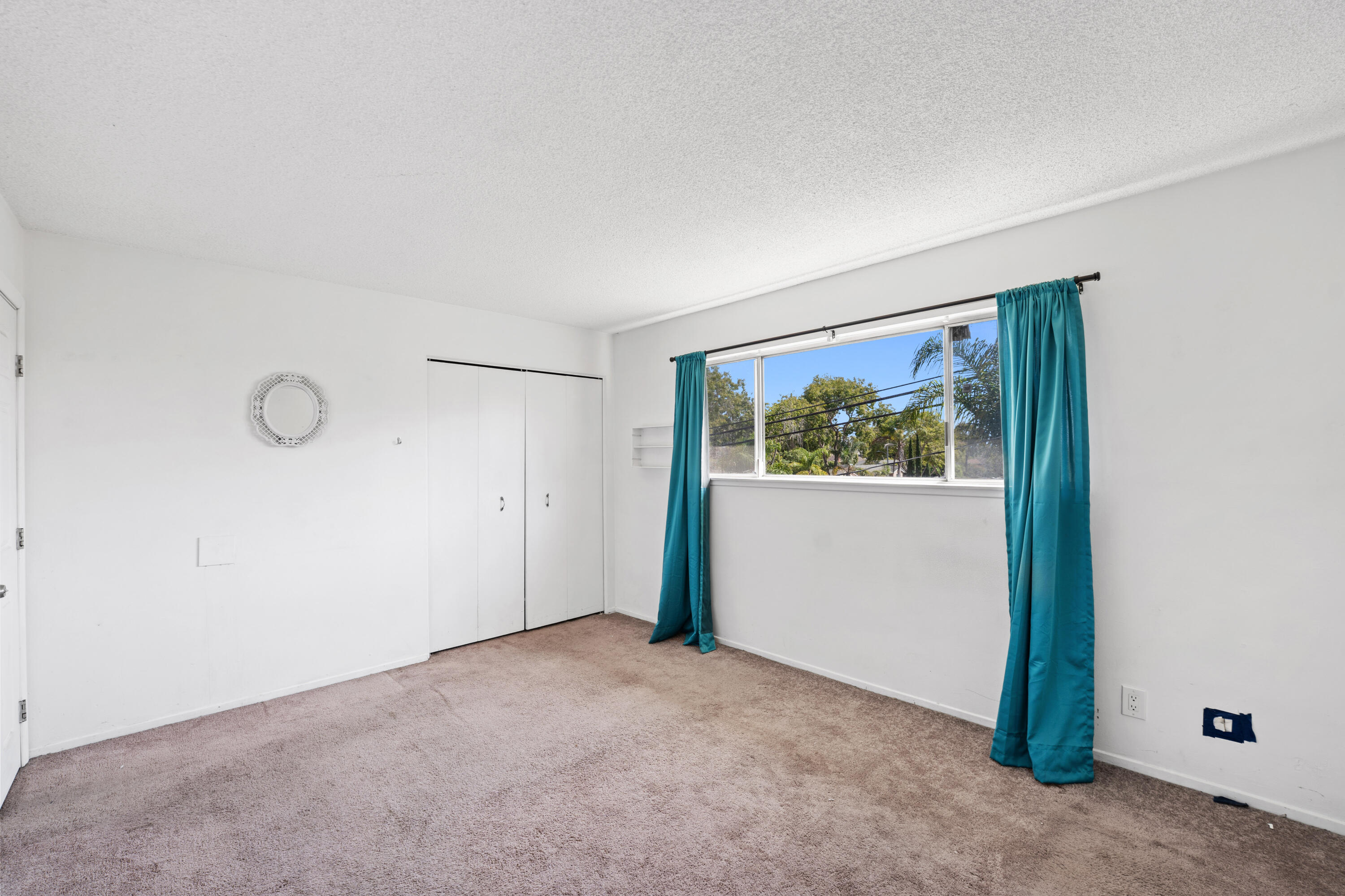Empty bedroom with beige carpet, built-in closet, and a wide window framed by teal curtains.