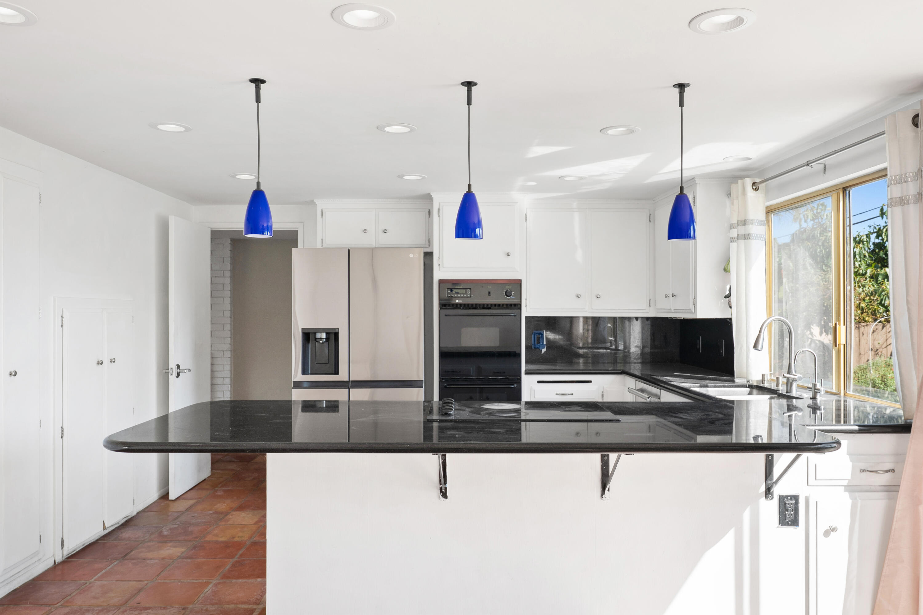 Bright white kitchen with black stone peninsula, blue pendant lights, stainless steel appliances, and terracotta tile floor.