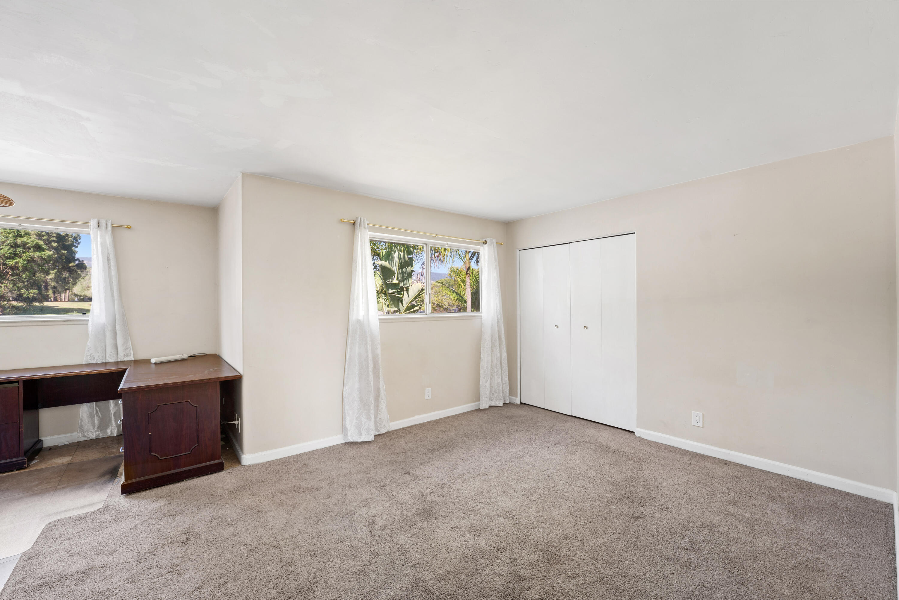 Bedroom with beige carpet, a built-in closet, and curtained windows beside a dark wood desk.