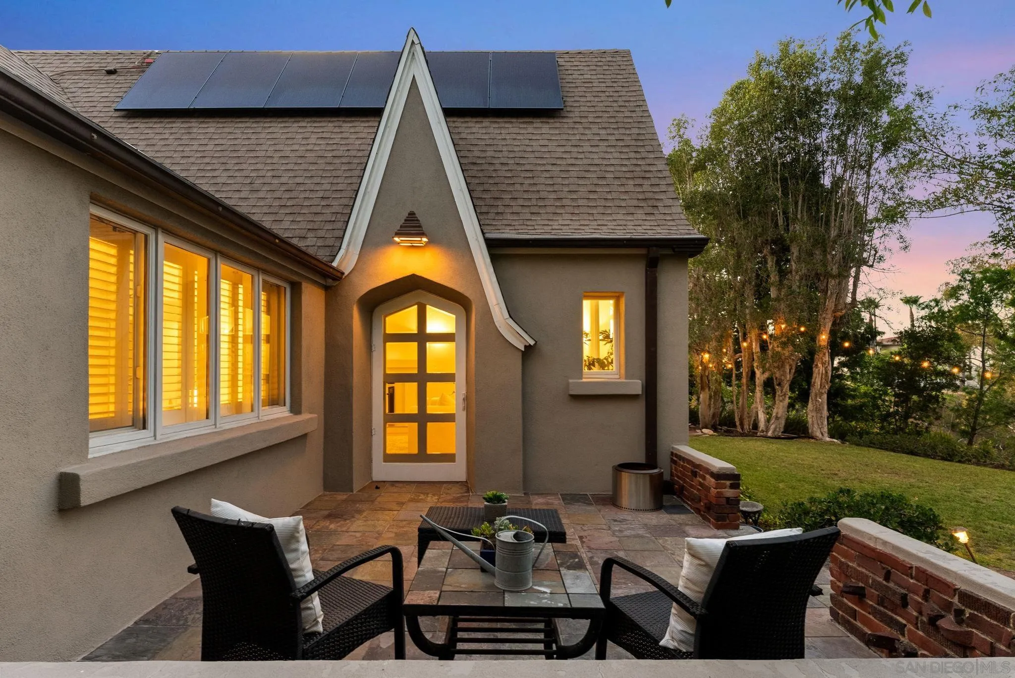 Home patio with black outdoor seating, solar panels, and warmly lit windows at dusk.