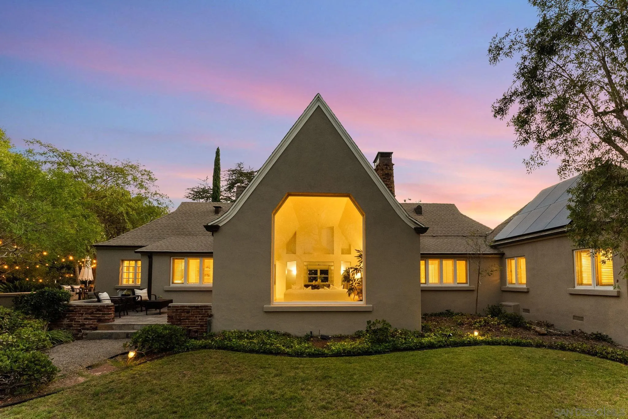 Gray house at dusk with a dramatic gable window, solar panels, and landscaped patio.