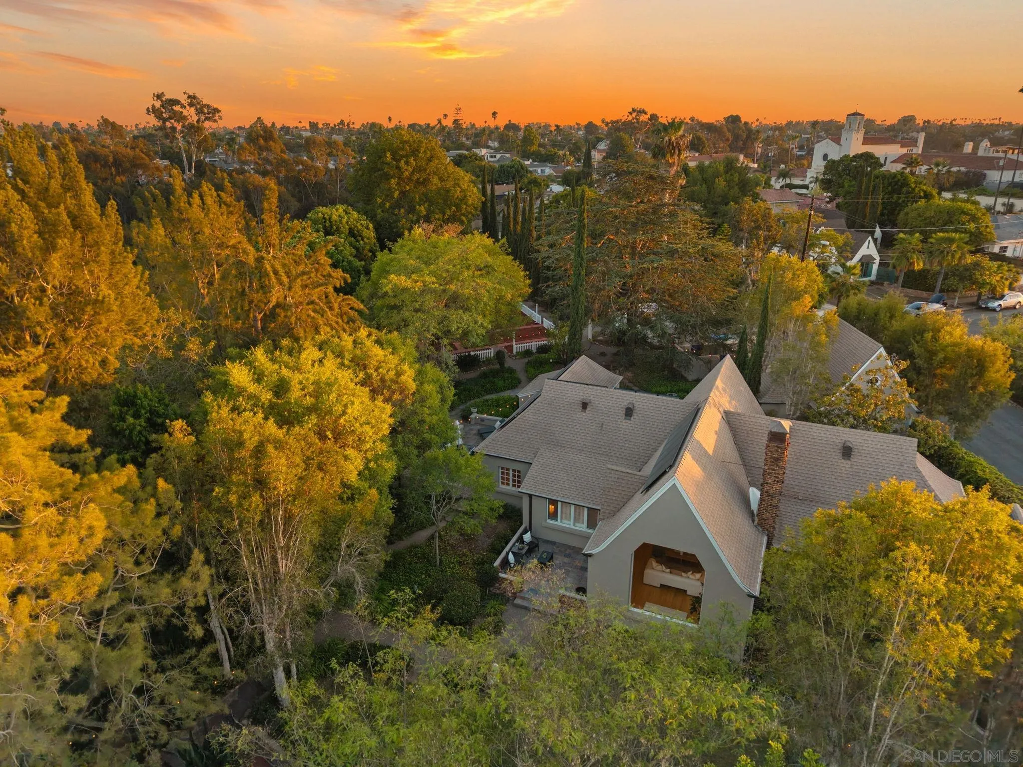 Aerial view of a gray-roofed home nestled among mature trees, with neighborhood homes beyond at sunset.