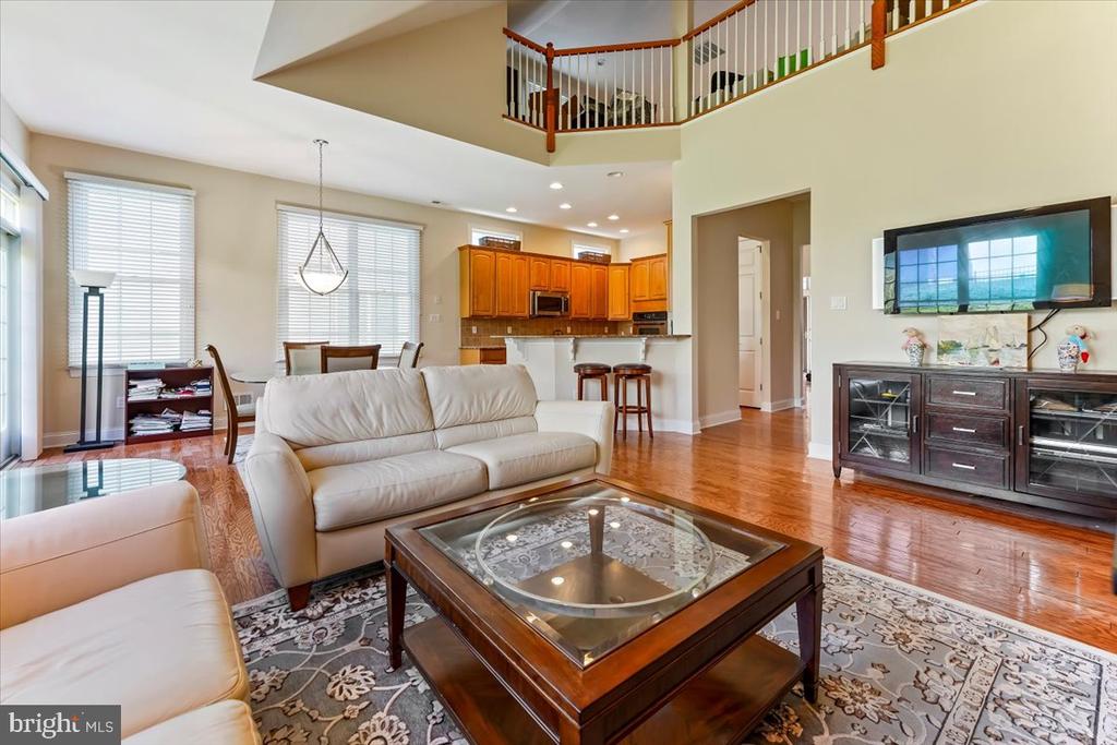 Open living room with white sofas, hardwood floors, and an adjoining kitchen beneath an upper-level railing.
