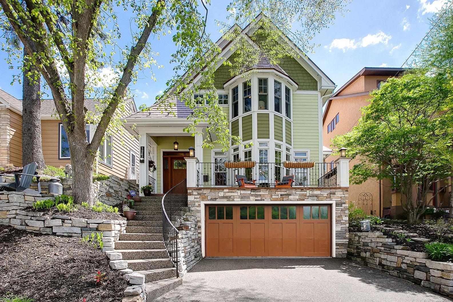 Green multi-story home with a bay window, stone retaining walls, front patio, and attached garage.