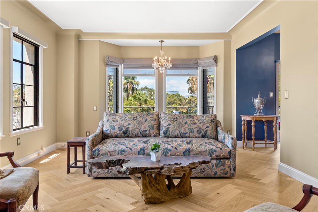 Living room with a patterned sofa, rustic coffee table, chandelier, and large windows overlooking greenery.