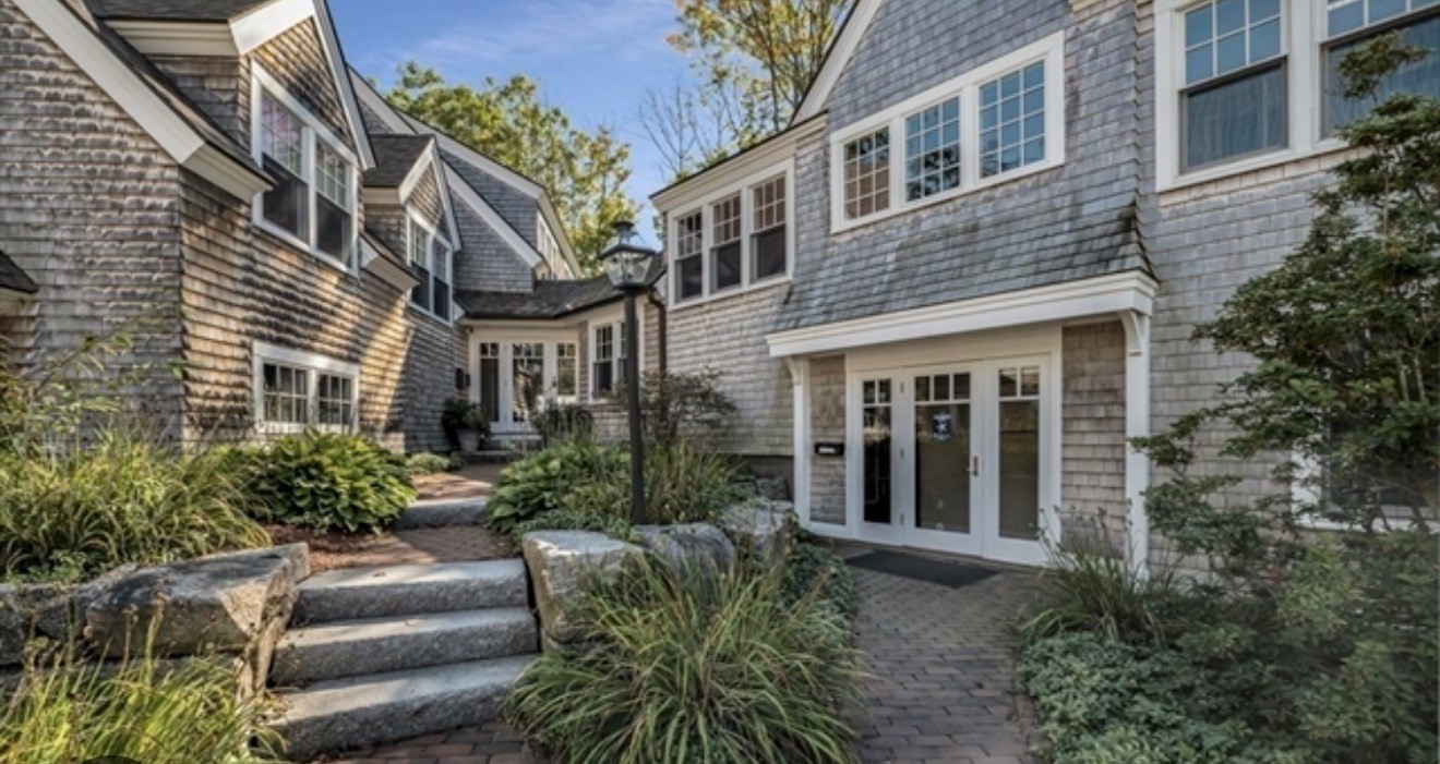 Gray-shingle house entrance with white-trimmed windows, stone steps, and a landscaped brick path.