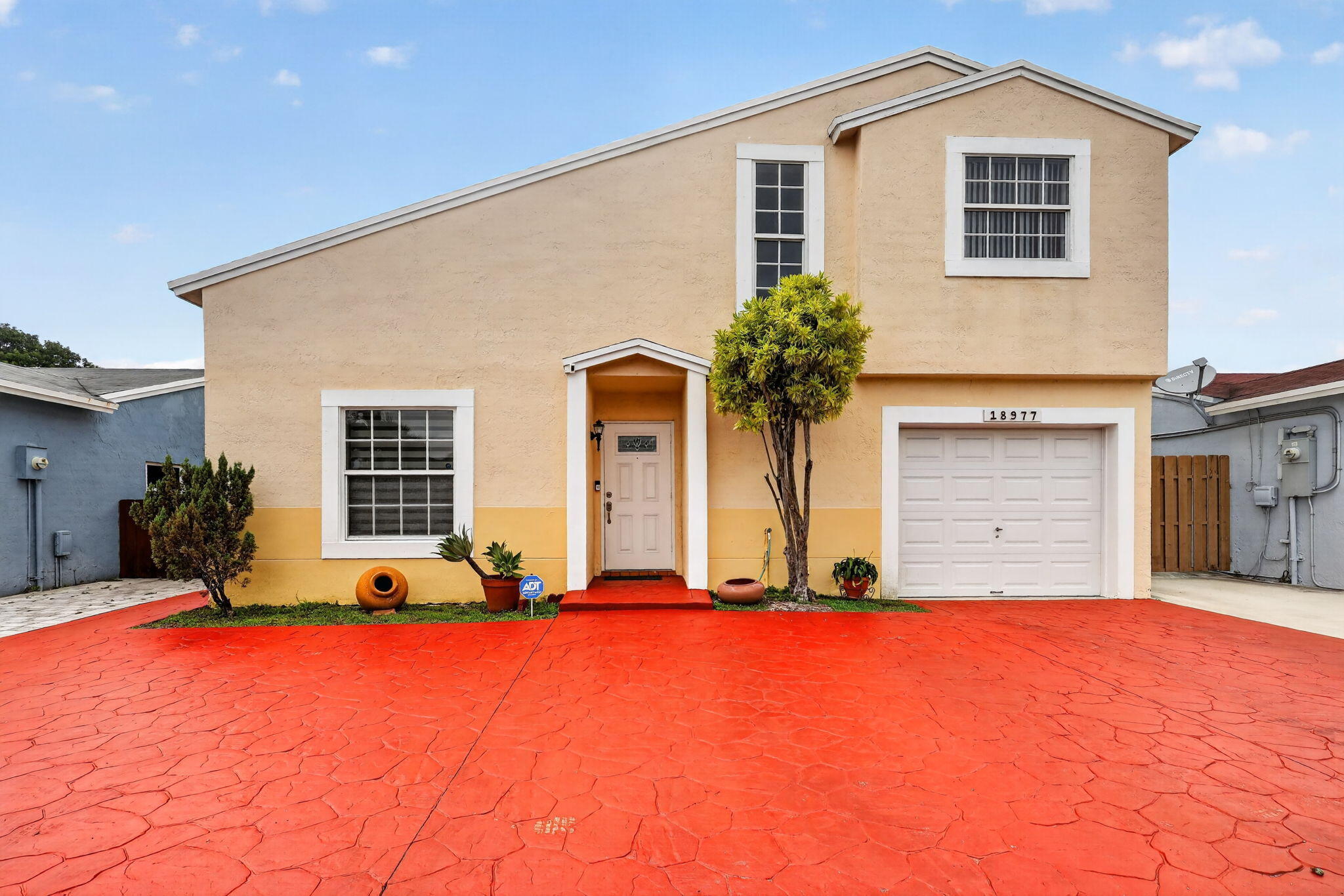 Two-story beige house with white trim, a garage, and a wide red stamped-concrete driveway.