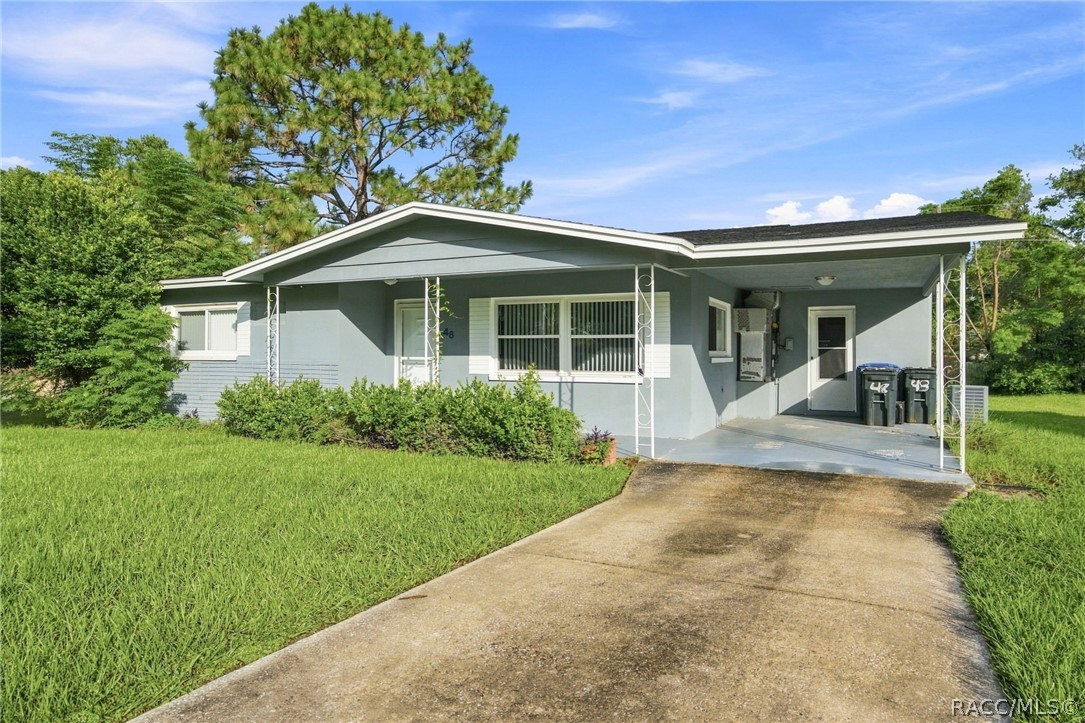 Single-story blue-gray house with a covered carport, concrete driveway, and mature trees.