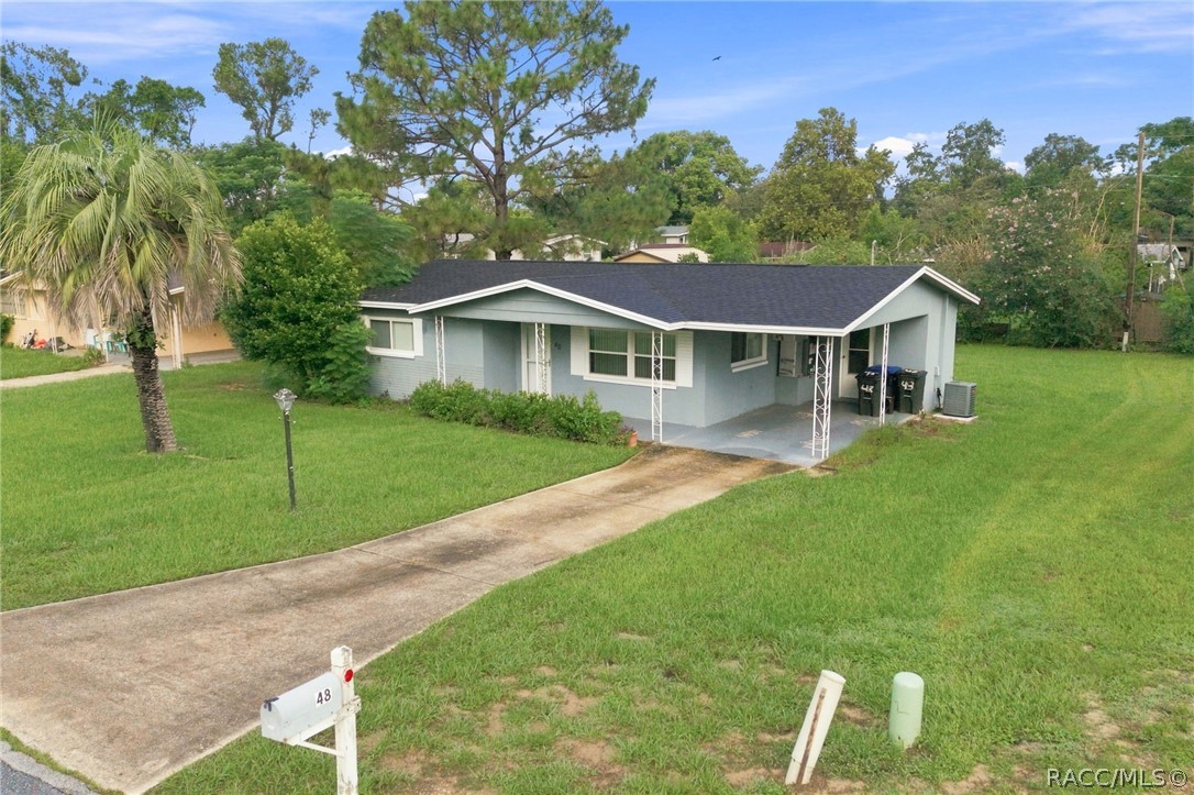 Single-story blue-gray house with a covered carport, broad lawn, palm tree, and curved driveway.