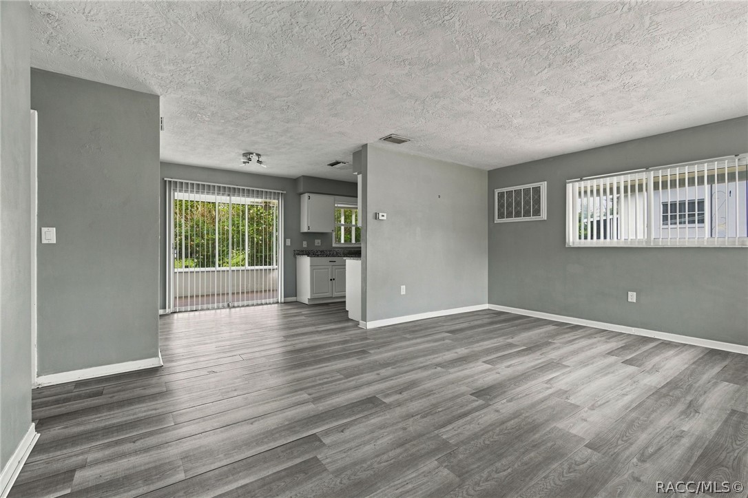 Empty living area with gray plank flooring, a small kitchen, and a sliding glass door.
