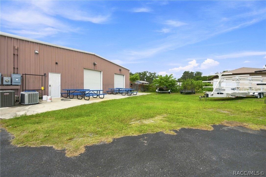 Commercial building with brown metal siding, roll-up doors, picnic tables, and a white trailer beside a grassy lot.
