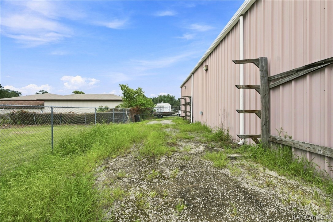 Pale pink metal-sided building beside a gravel lane, overgrown grass, and chain-link fence.