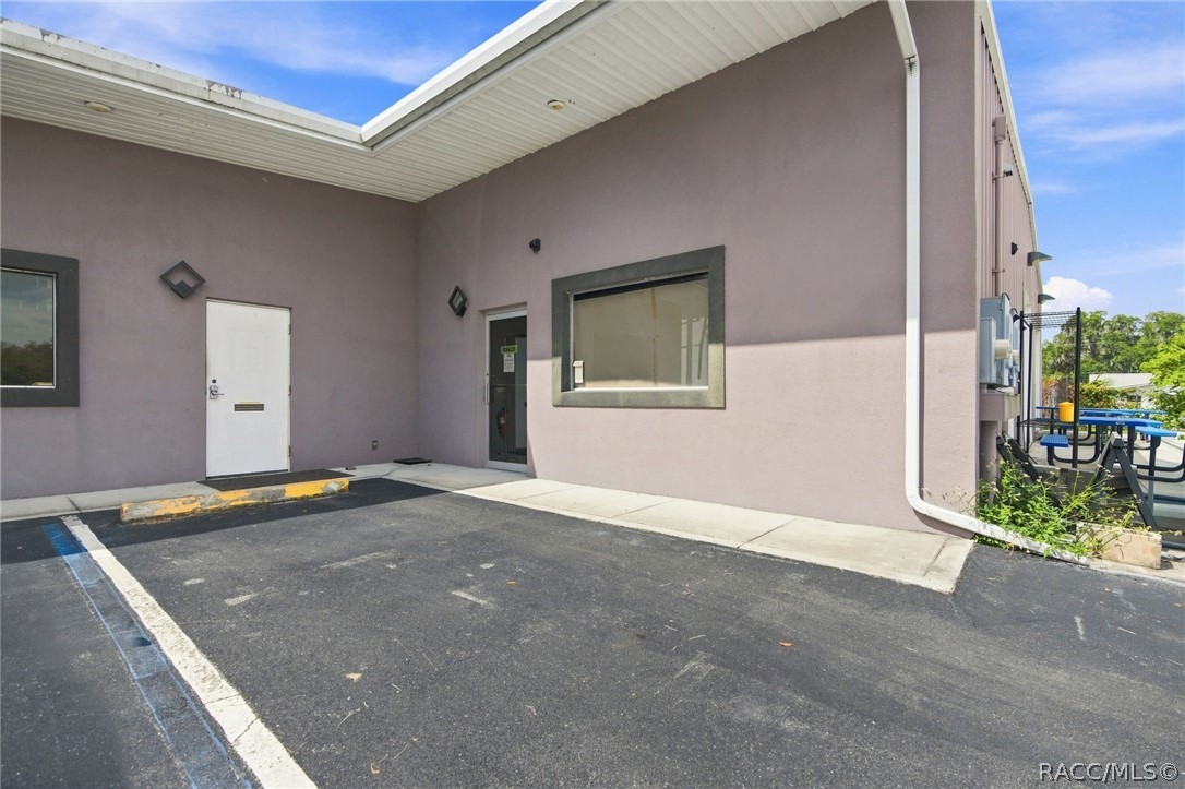 Mauve building exterior with a covered entrance, large window, and picnic tables beside the parking area.