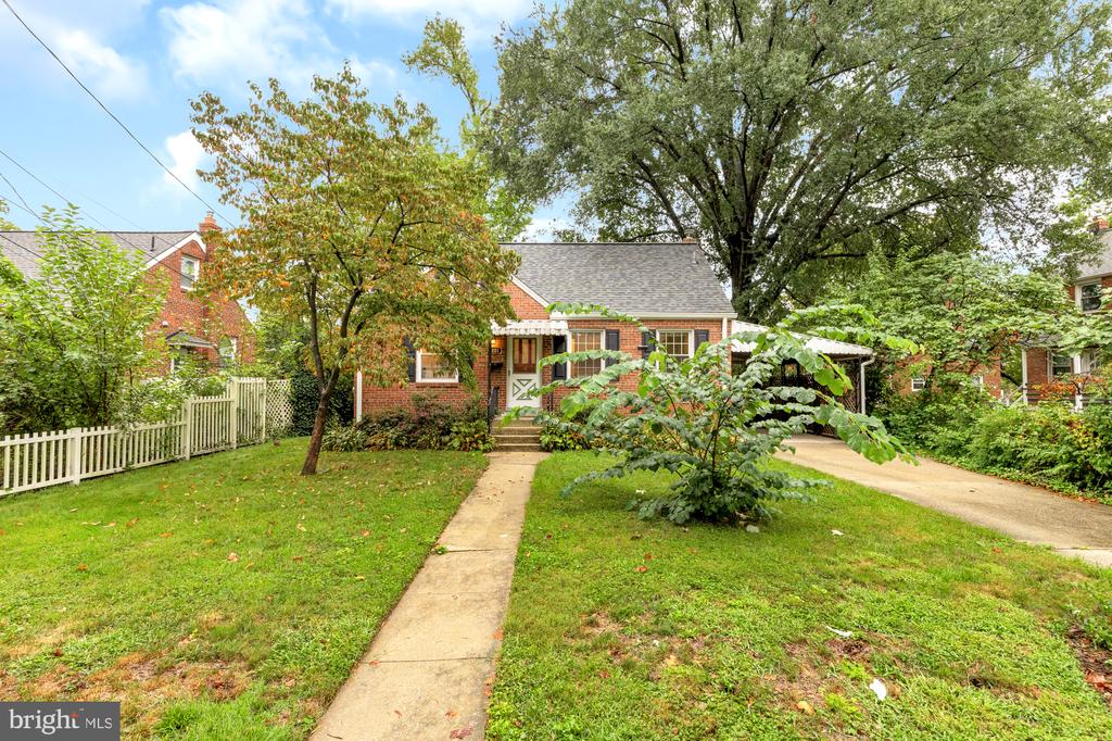 Red-brick house with a covered entrance, concrete walkway, and mature trees.