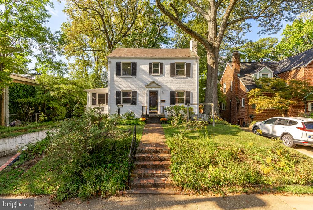 Two-story white house with dark shutters, brick front steps, a landscaped yard, and mature shade trees.
