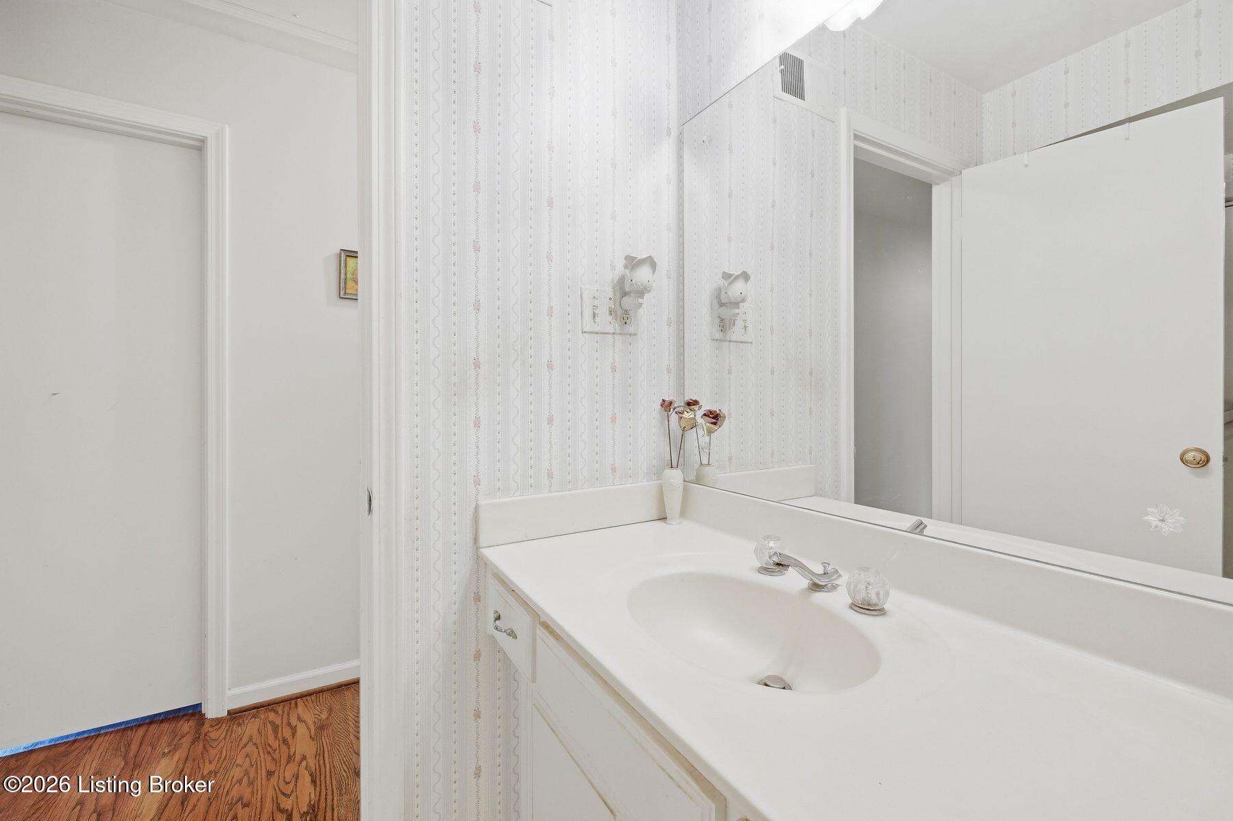 Bathroom vanity with a large mirror, patterned wallpaper, and an open doorway to a wood-floored room.