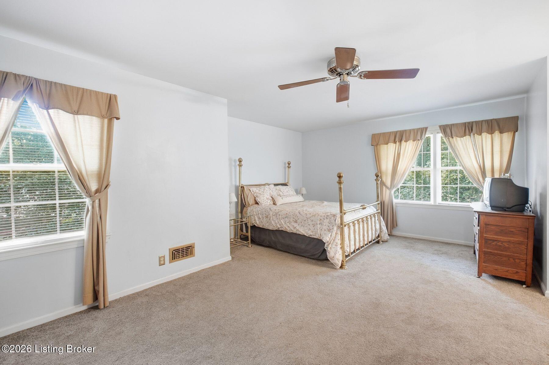 Bedroom with a brass four-poster bed, beige carpet, and two large curtained windows.