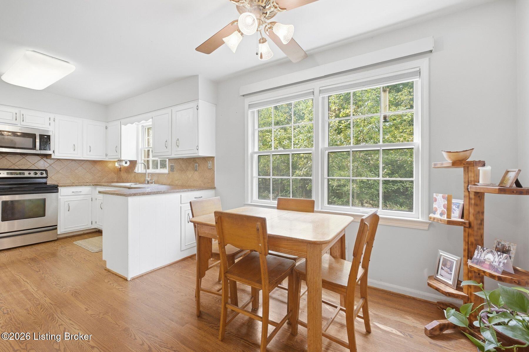 Bright kitchen and dining area with white cabinets, hardwood floors, and large windows overlooking greenery.