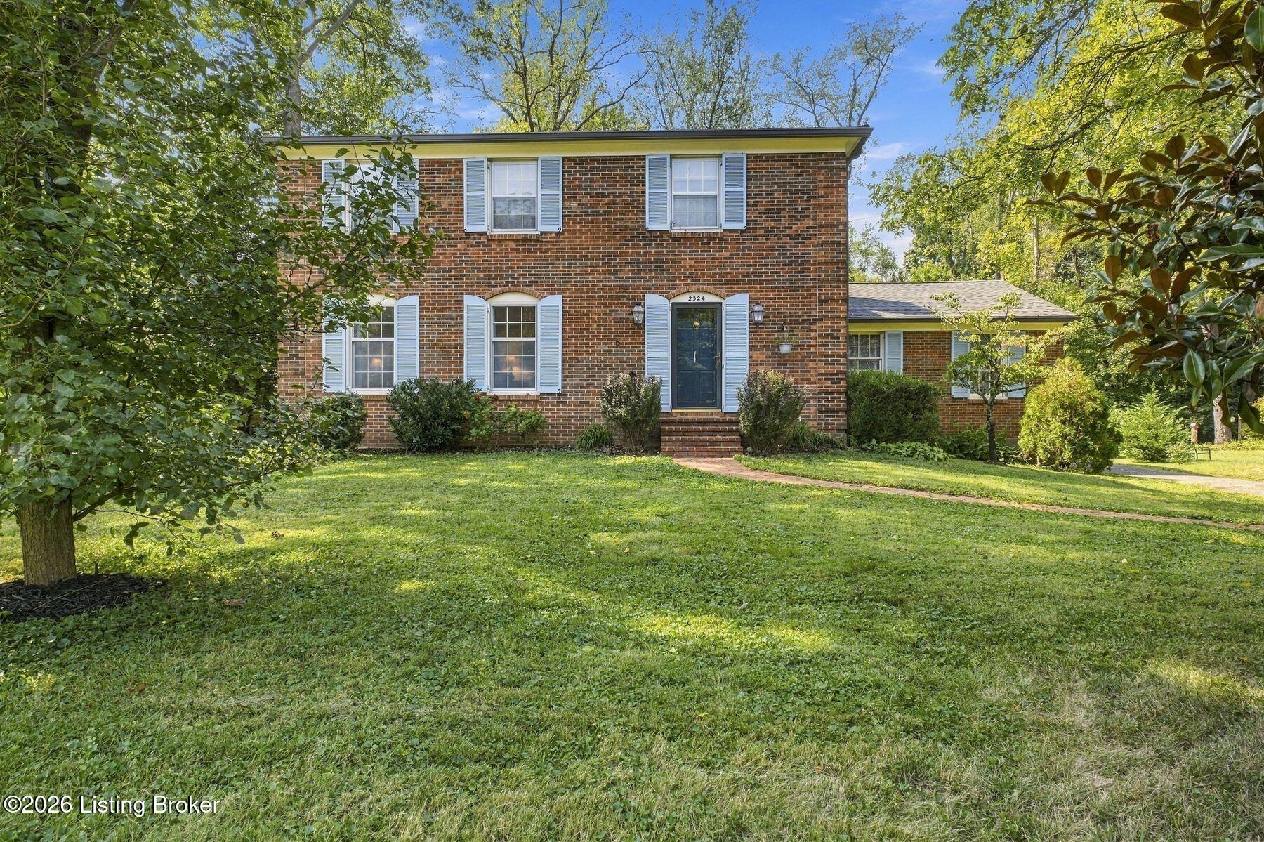 Two-story brick house with white shutters and blue front door, framed by mature trees and a broad lawn.