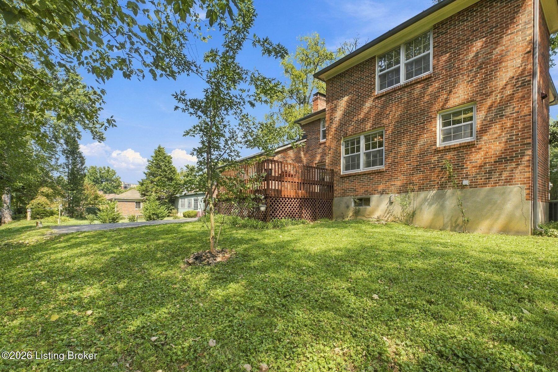 Two-story brick house with a raised wooden deck overlooking a leafy lawn.