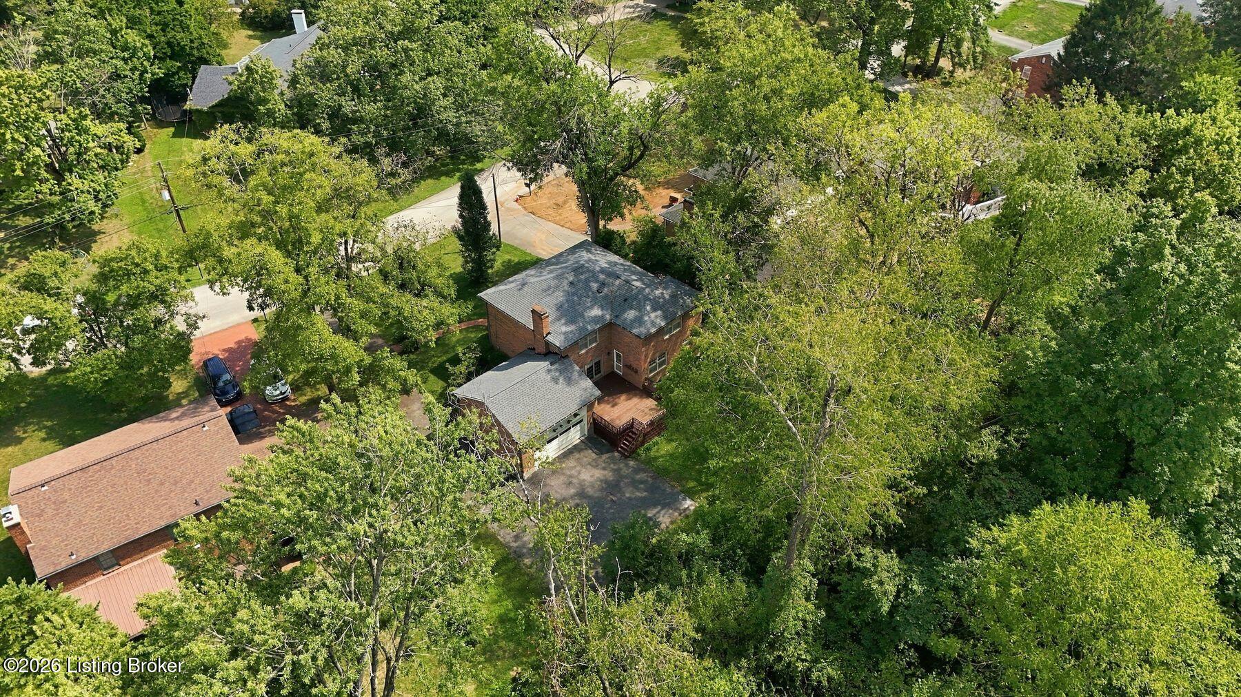 Aerial view of a two-story brick home surrounded by mature trees, with a driveway and rear deck.