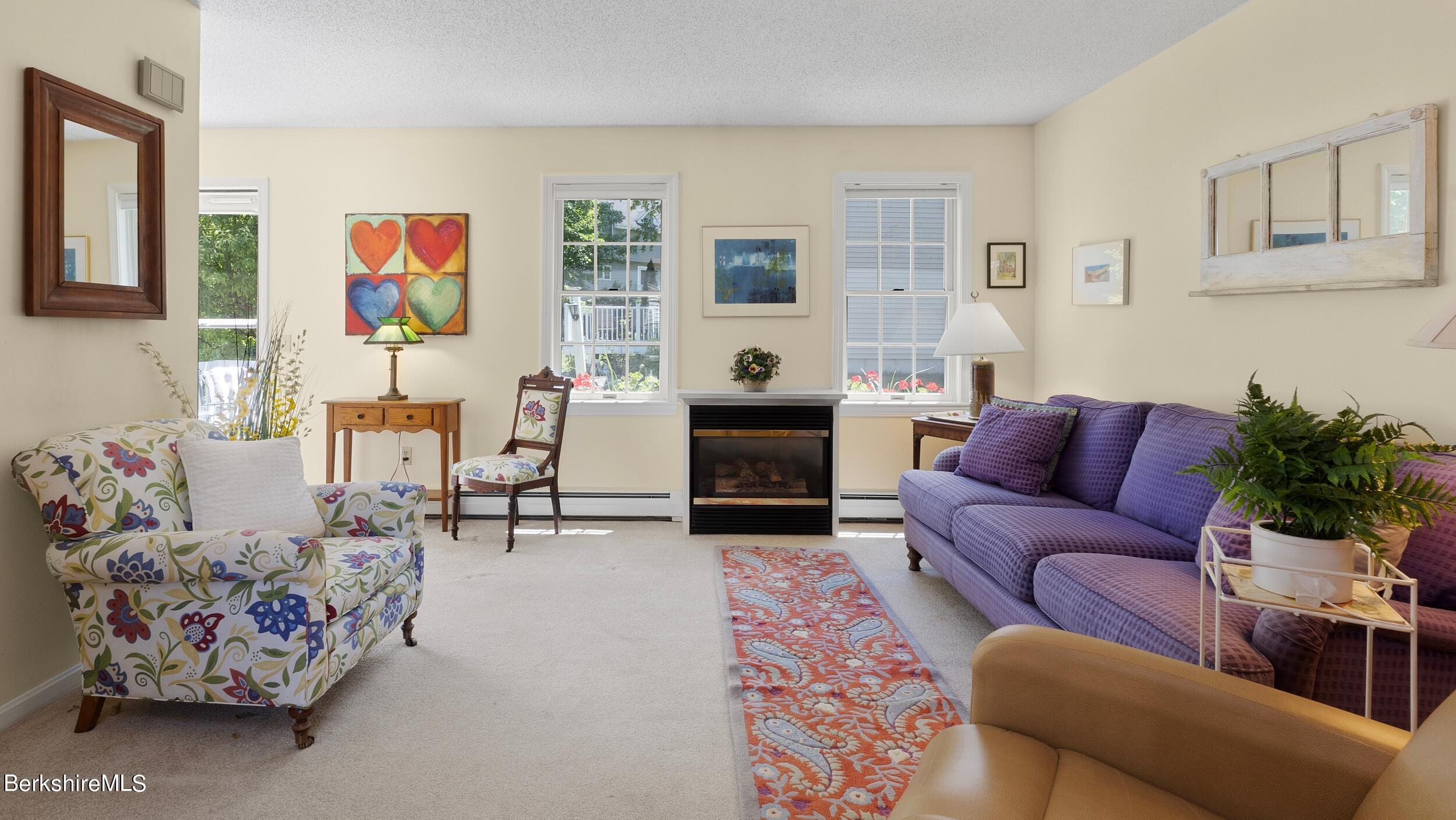 Living room with a central fireplace, purple sofa, floral armchair, and bright windows.