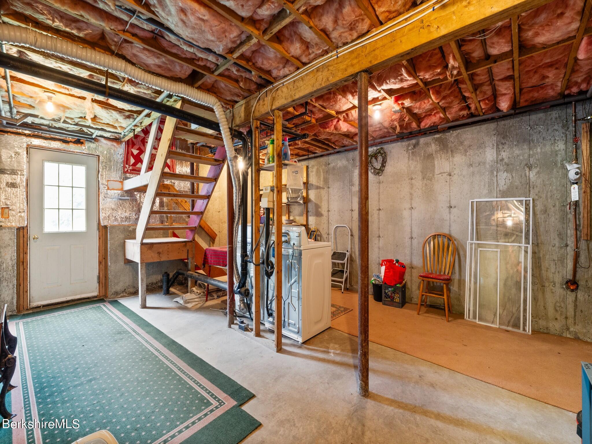 Unfinished basement with exposed insulation, wooden stairs, concrete walls, and laundry appliances.