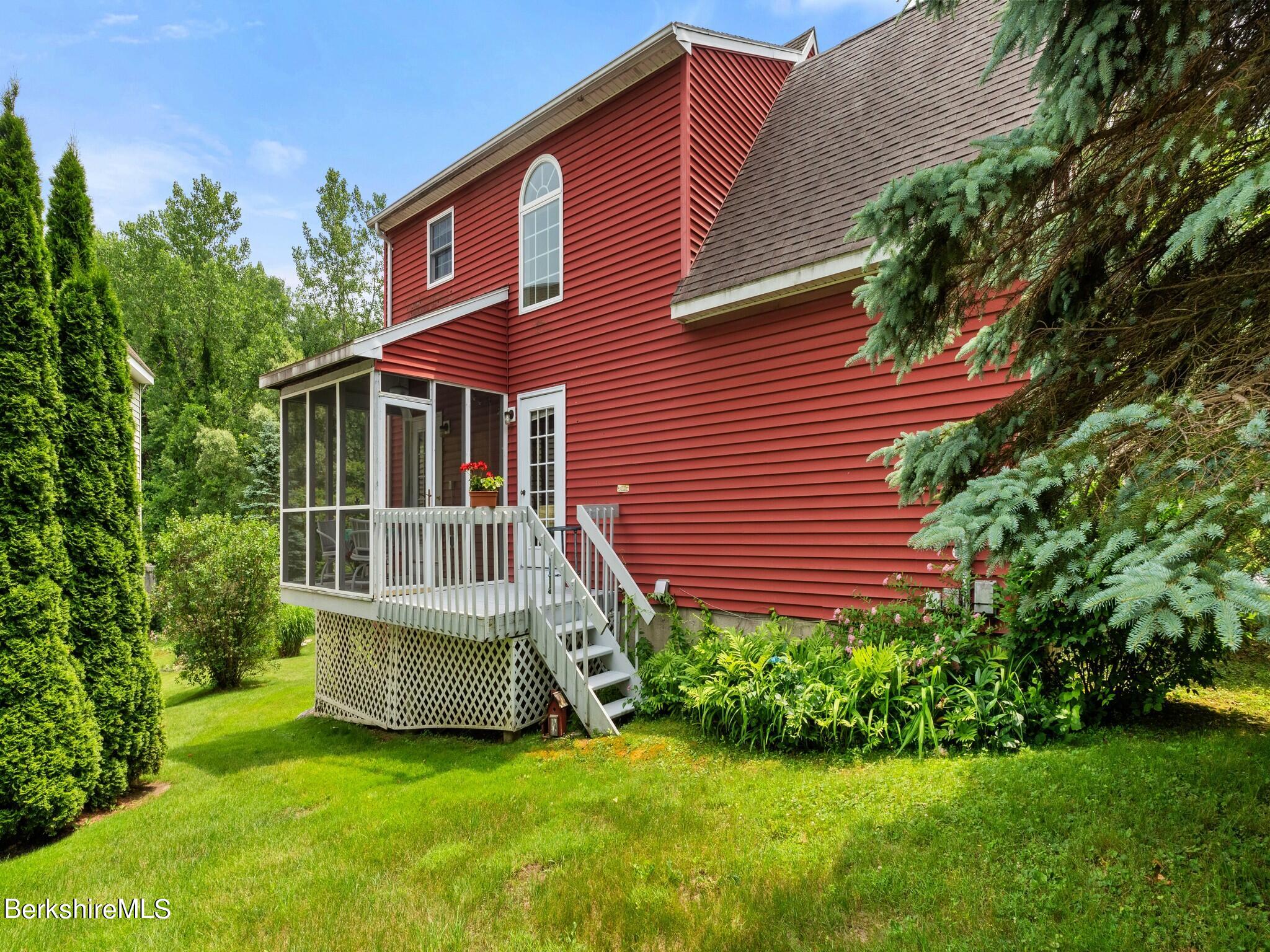 Red two-story house with a screened porch and elevated white deck amid mature trees and landscaped gardens.