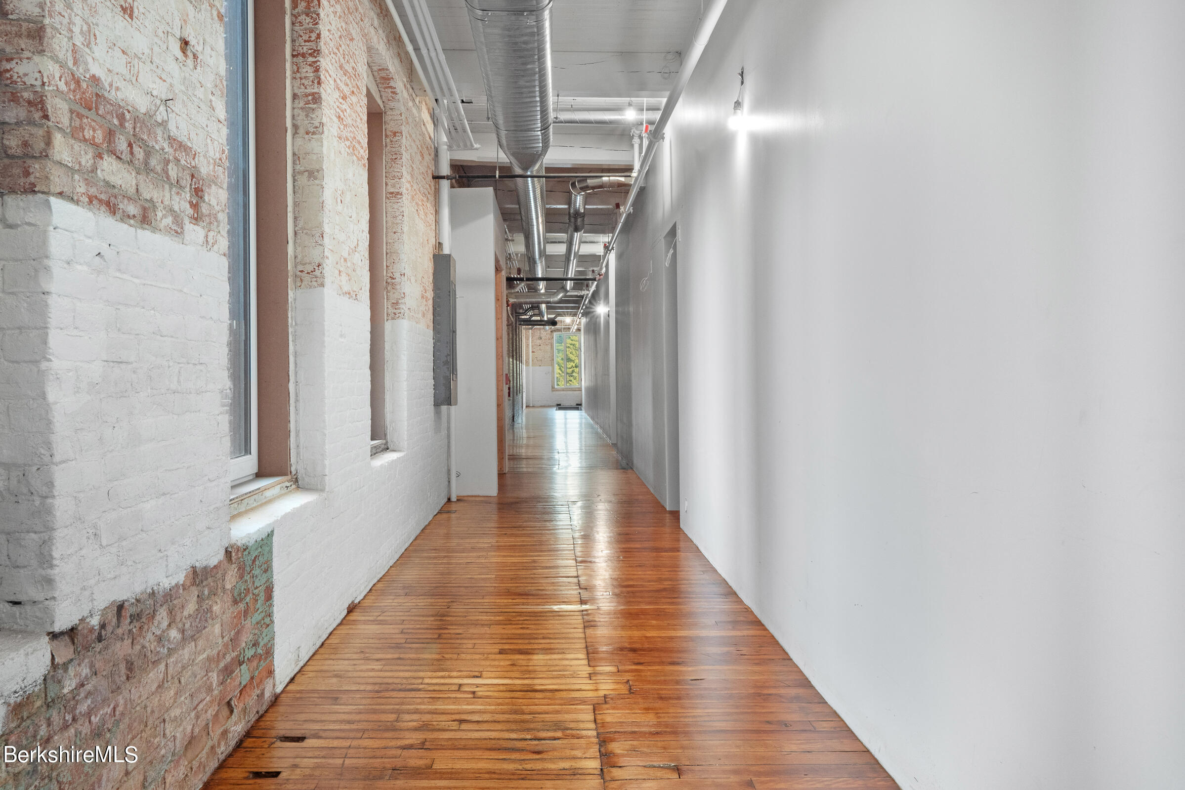 Vacant hallway with polished wood floors, white-painted brick, large windows, and exposed ceiling ducts.