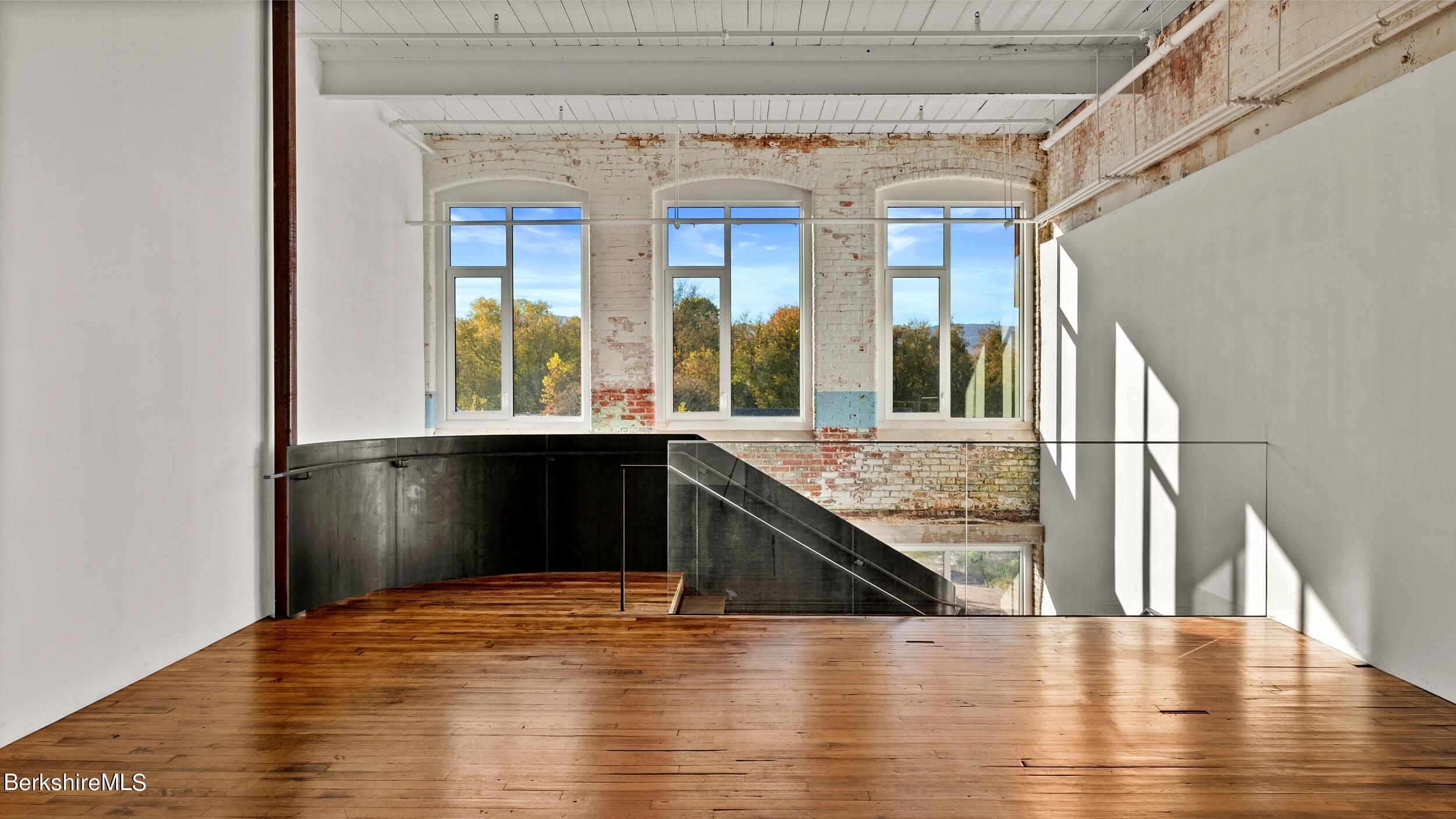 Vacant loft with wide plank wood floors, exposed brick, tall windows, and a glass-railed staircase.