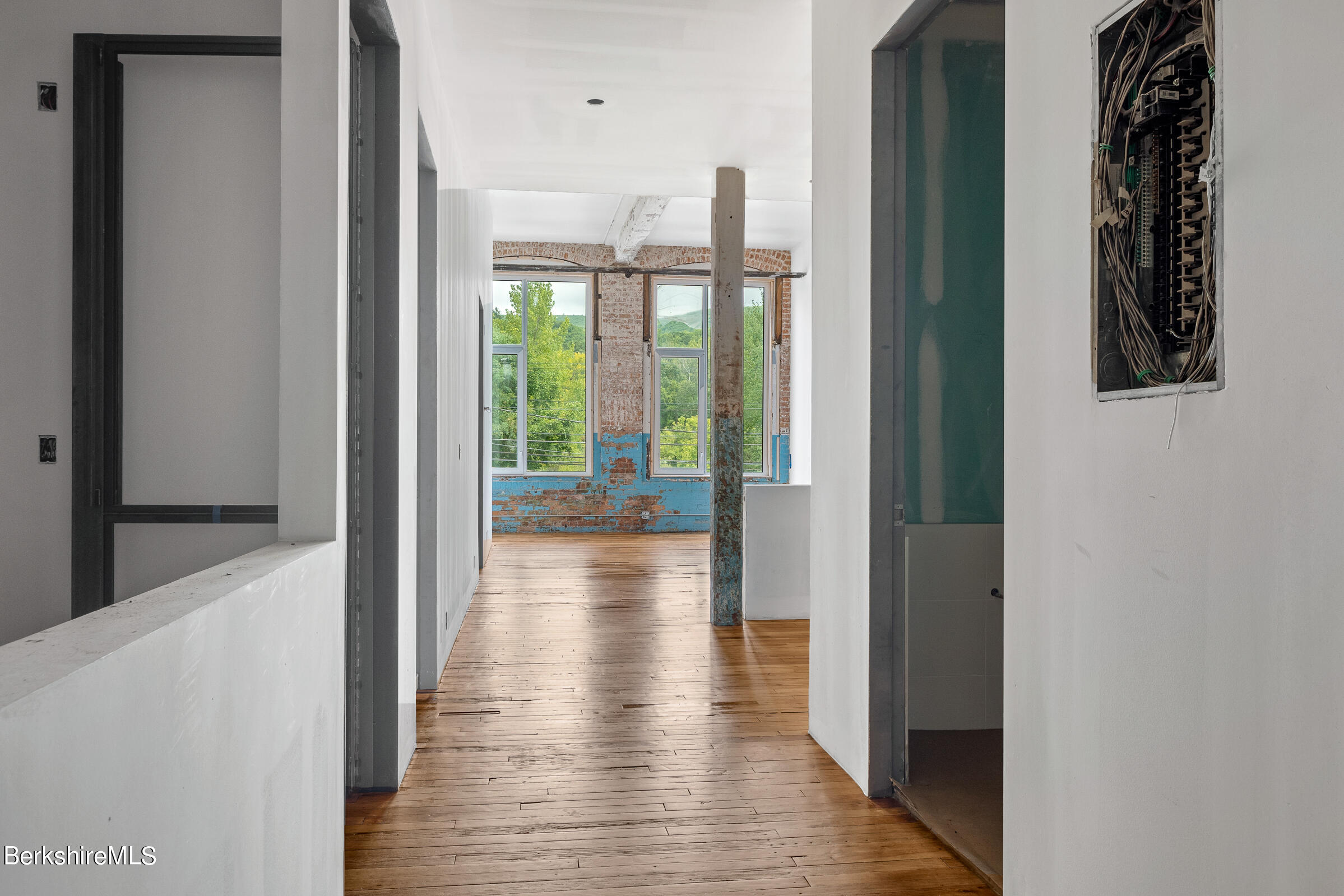 Unfinished hallway with exposed wood floors, a weathered support column, and tall windows overlooking green hills.