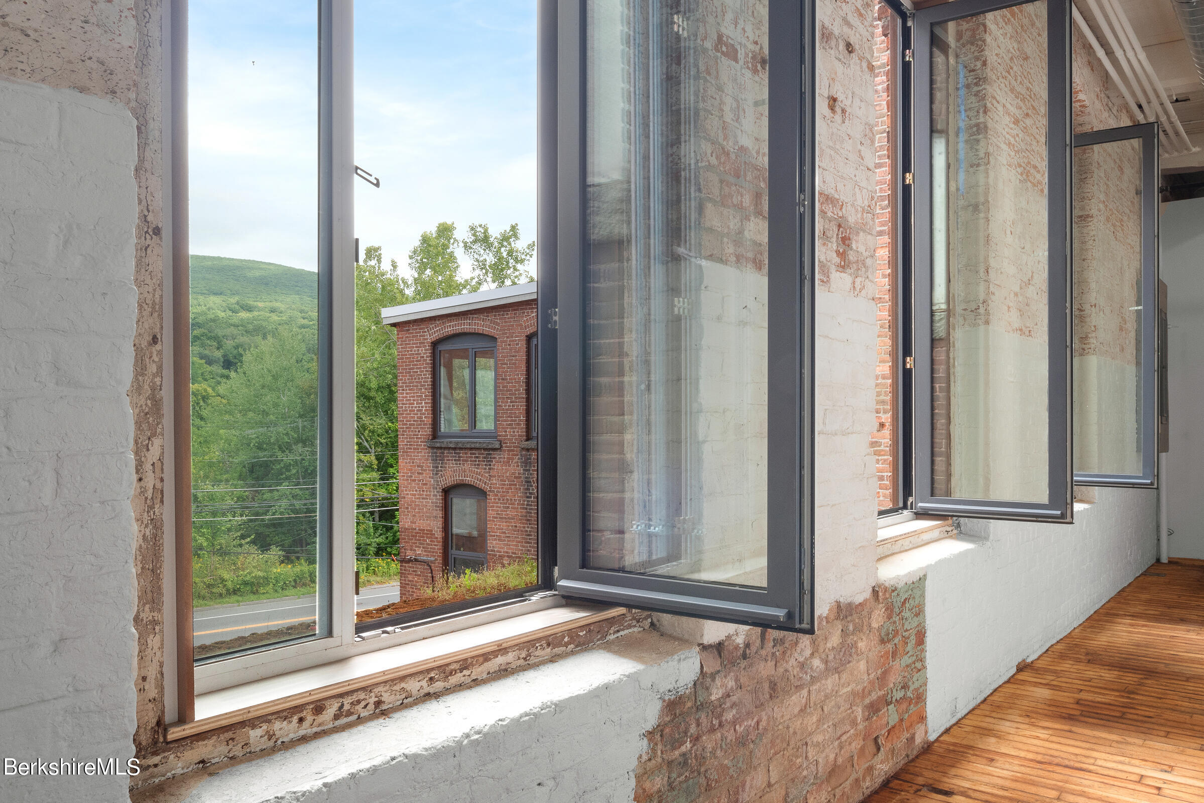Empty brick interior with tall open windows, hardwood floors, and a view of a wooded hillside.