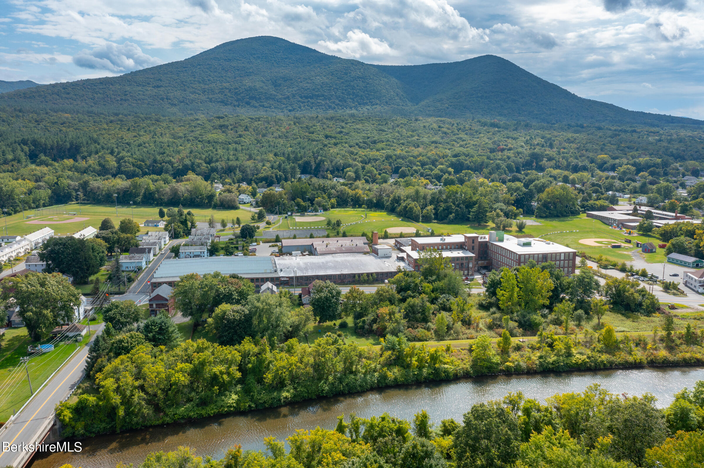 Aerial view of a brick complex beside a river, with ballfields and wooded mountains beyond.