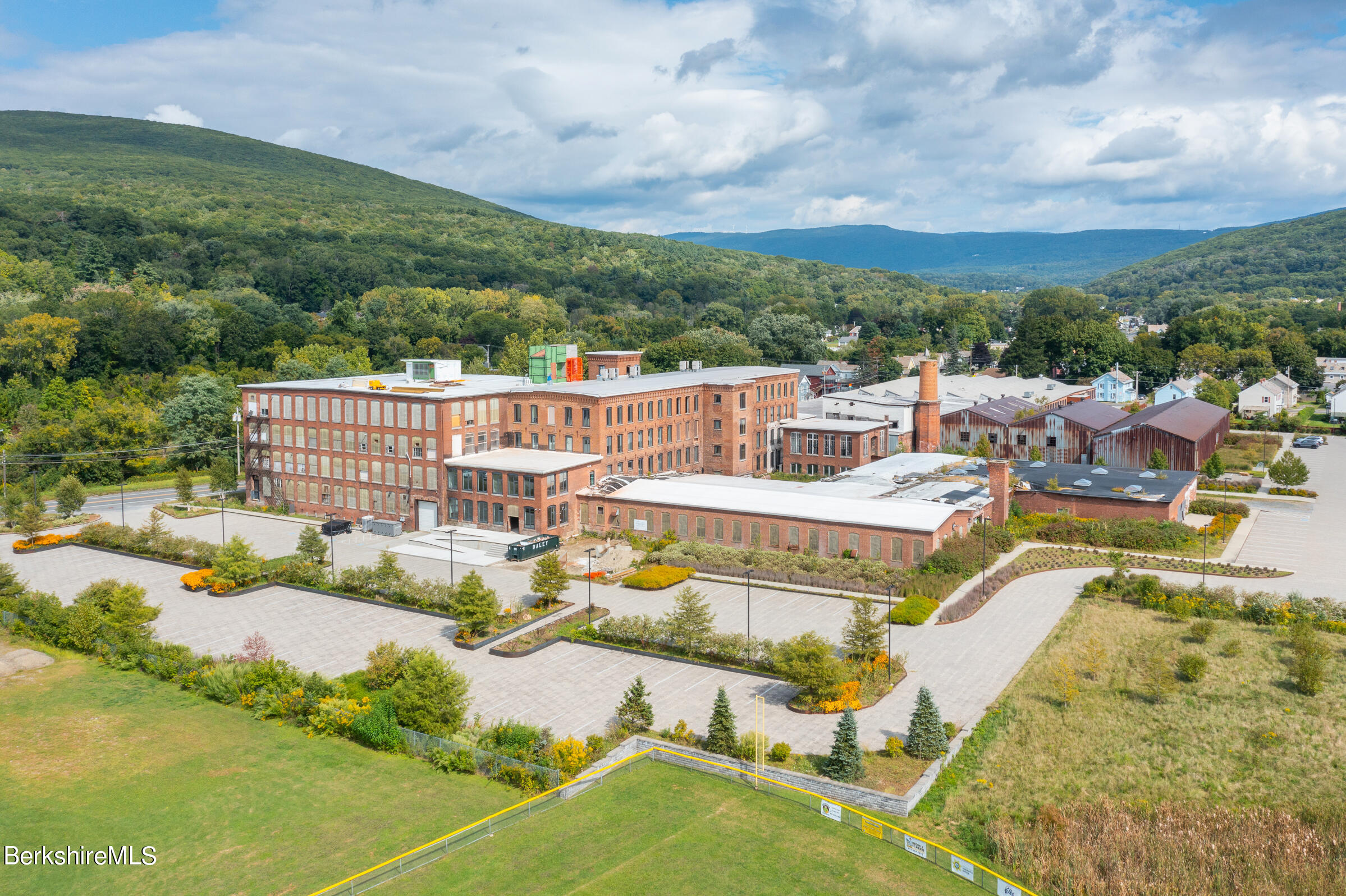 Aerial view of a brick mill complex and parking lot, with wooded hills in the background.