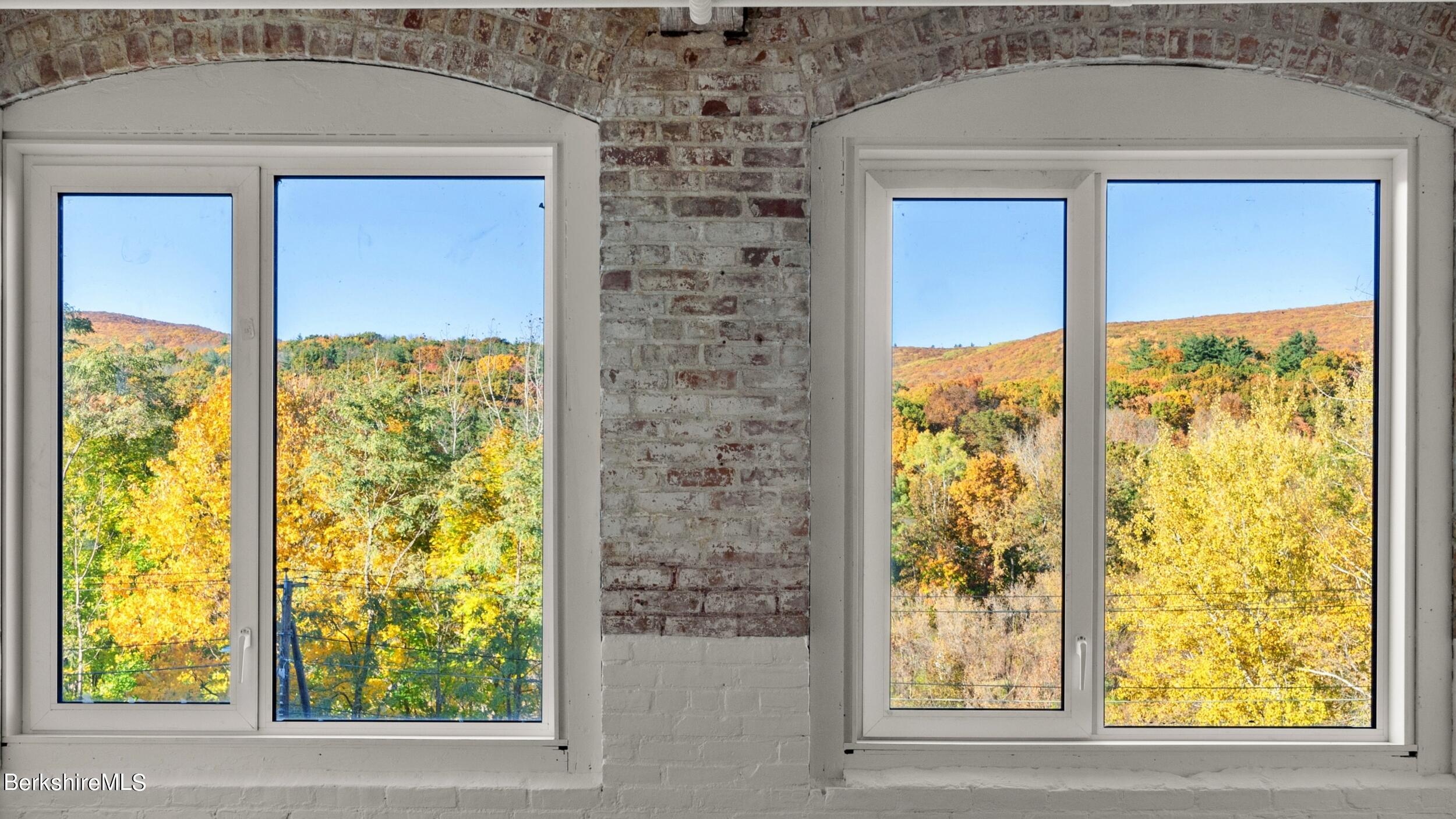 Two arched windows frame an autumn hillside view from a brick interior.