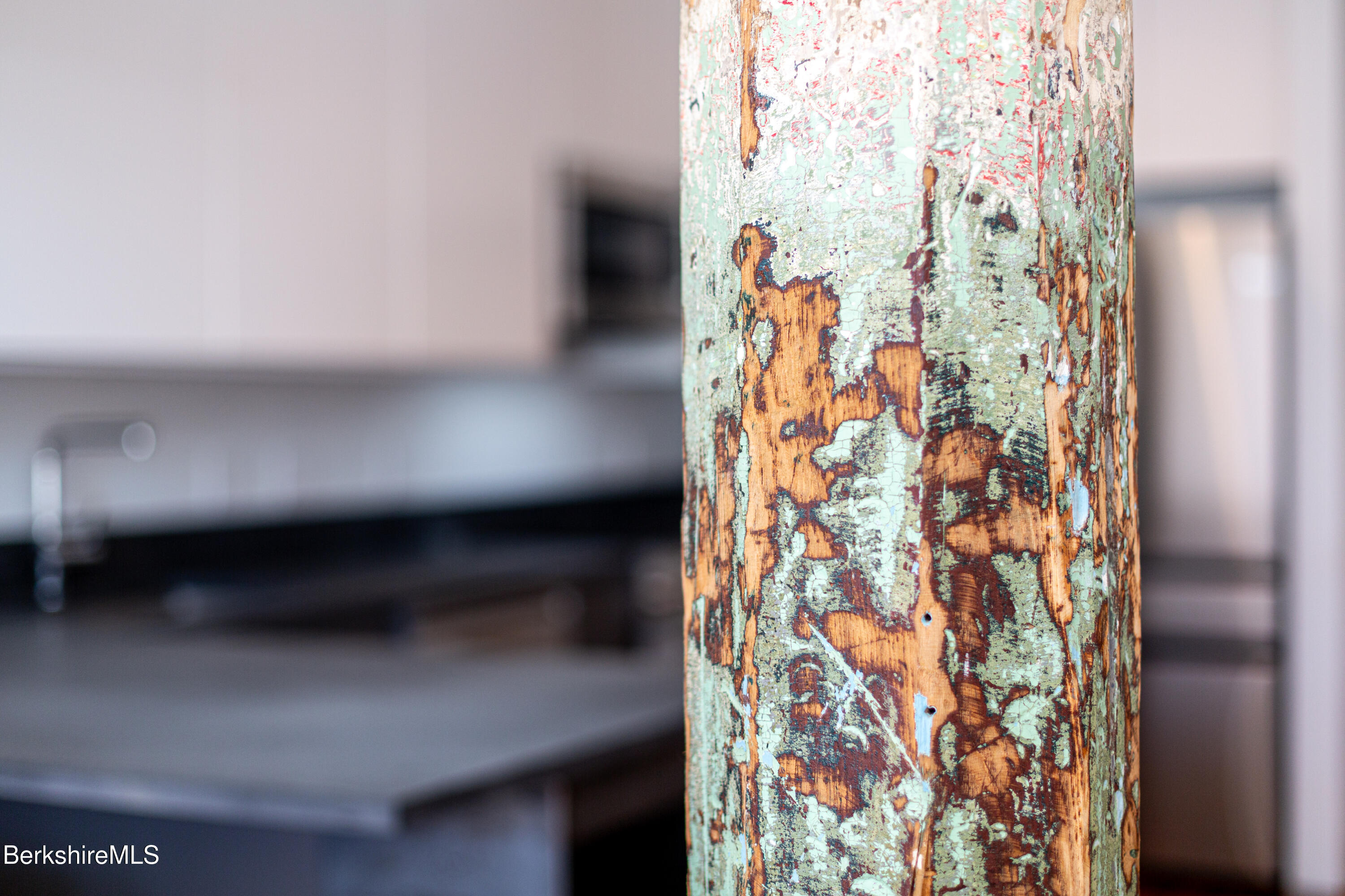 Distressed wooden post with chipped mint-green and orange paint, with a kitchen counter blurred behind it.