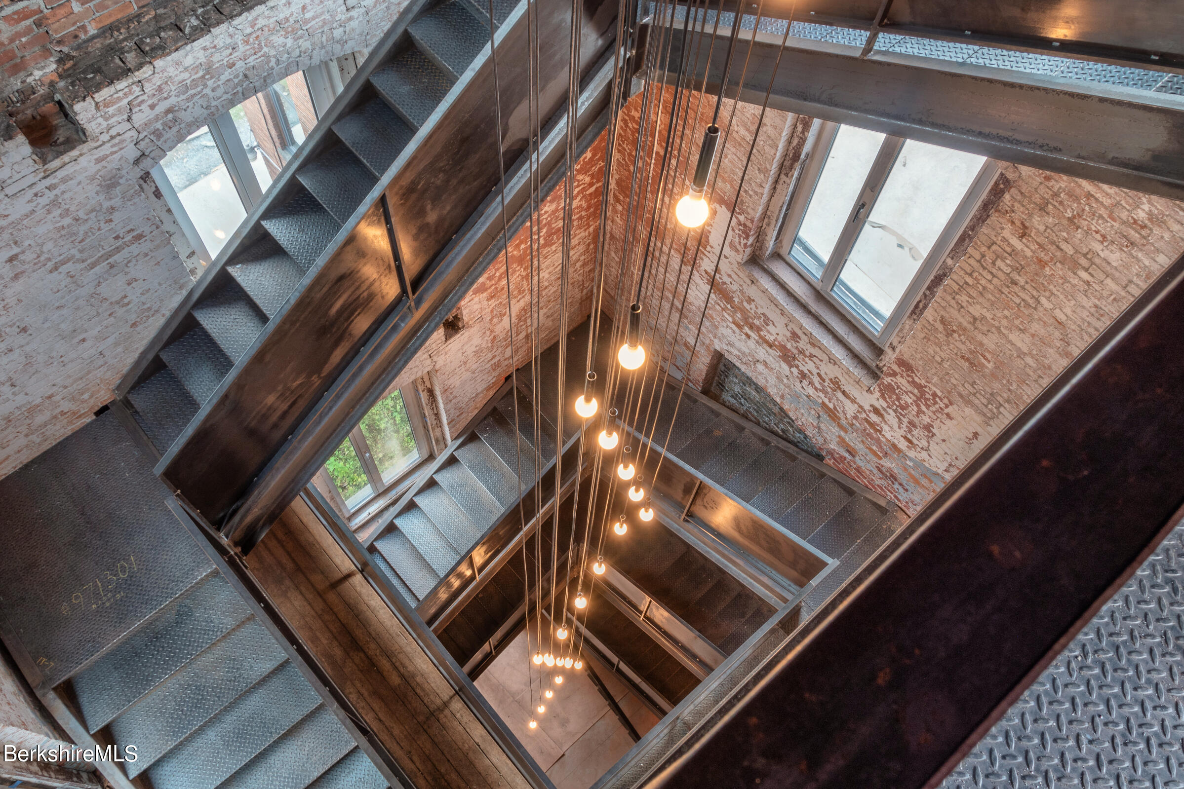 Multi-level metal stairwell with exposed brick walls, tall windows, and hanging pendant lights.
