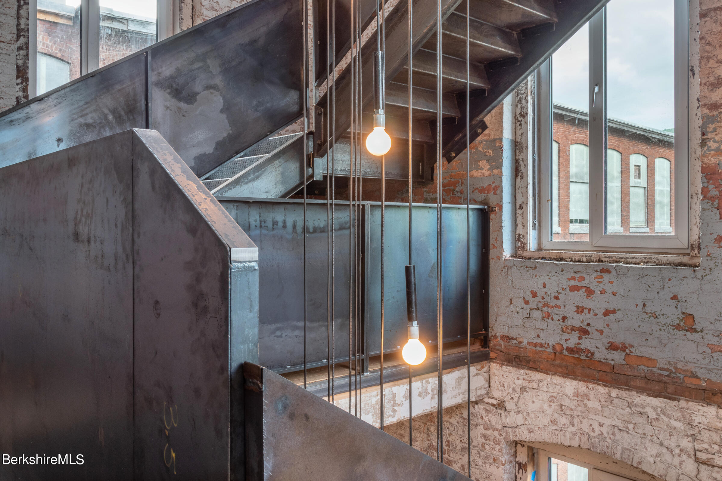 Industrial metal staircase with exposed brick walls, pendant bulbs, and tall windows.