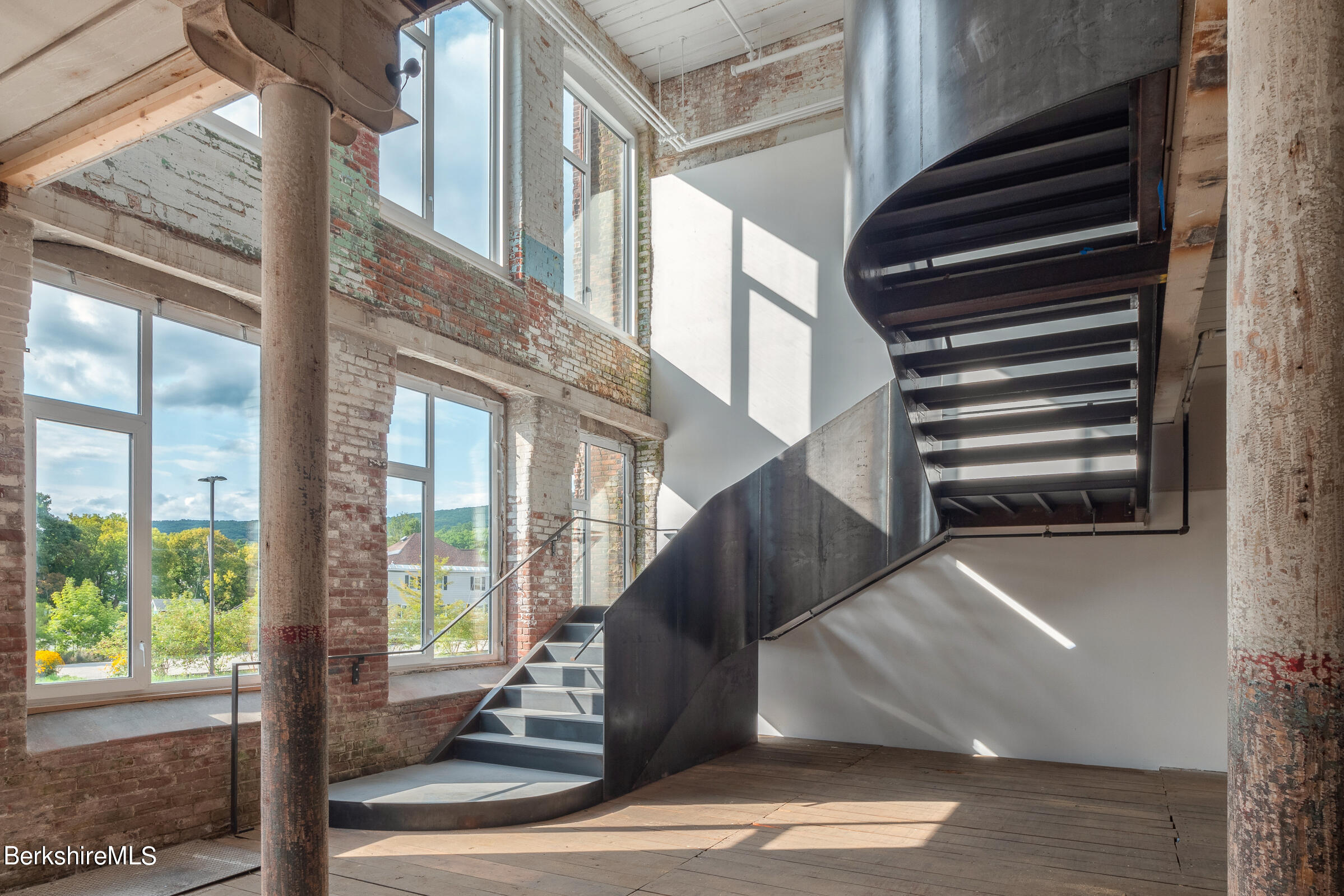 Industrial loft interior with exposed brick, tall windows, and a sculptural dark metal staircase.