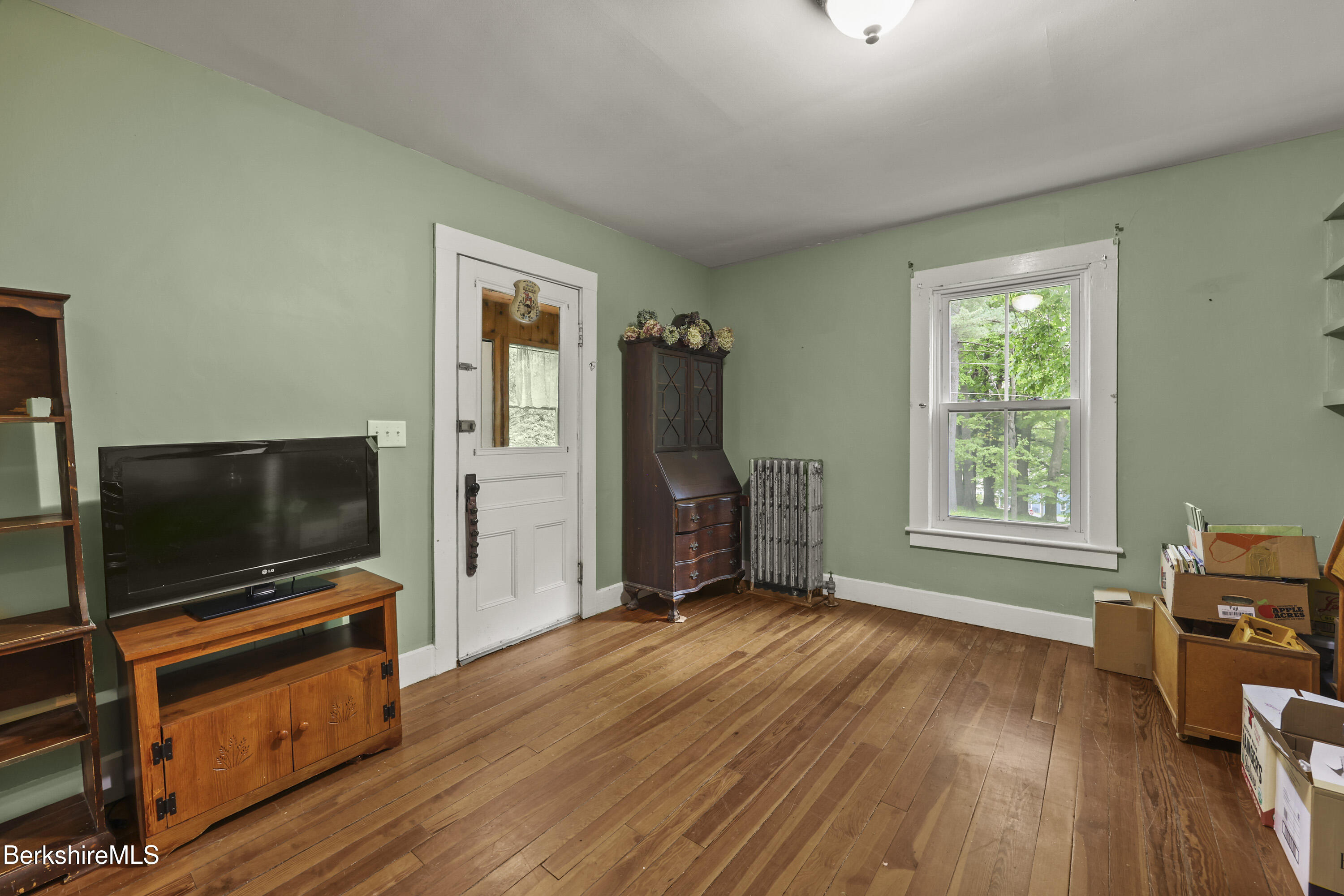 Green-walled room with hardwood floors, antique wooden furniture, a white door, and a window overlooking trees.