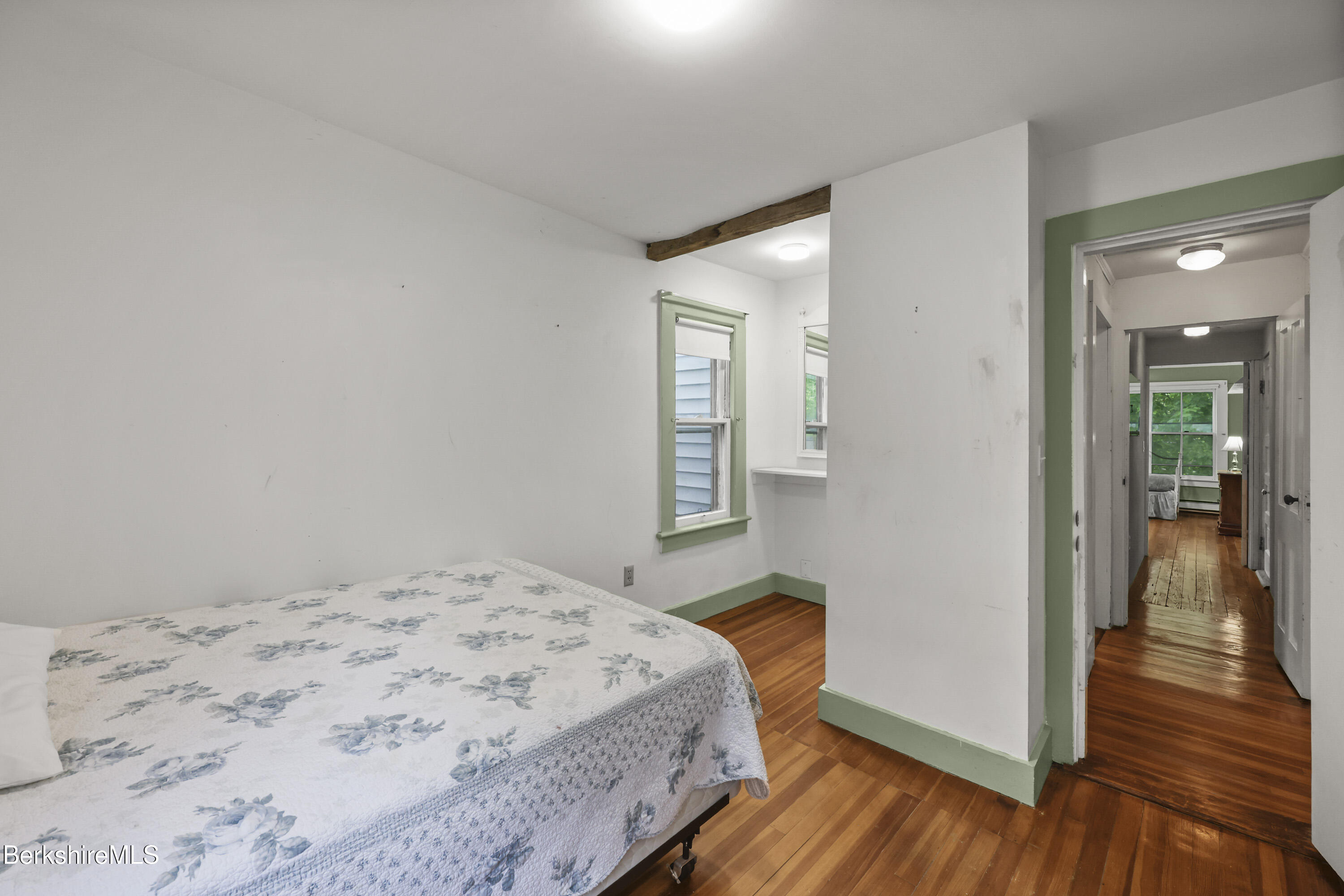 Bedroom with floral quilt, green-trimmed windows, exposed ceiling beam, and hardwood floors.