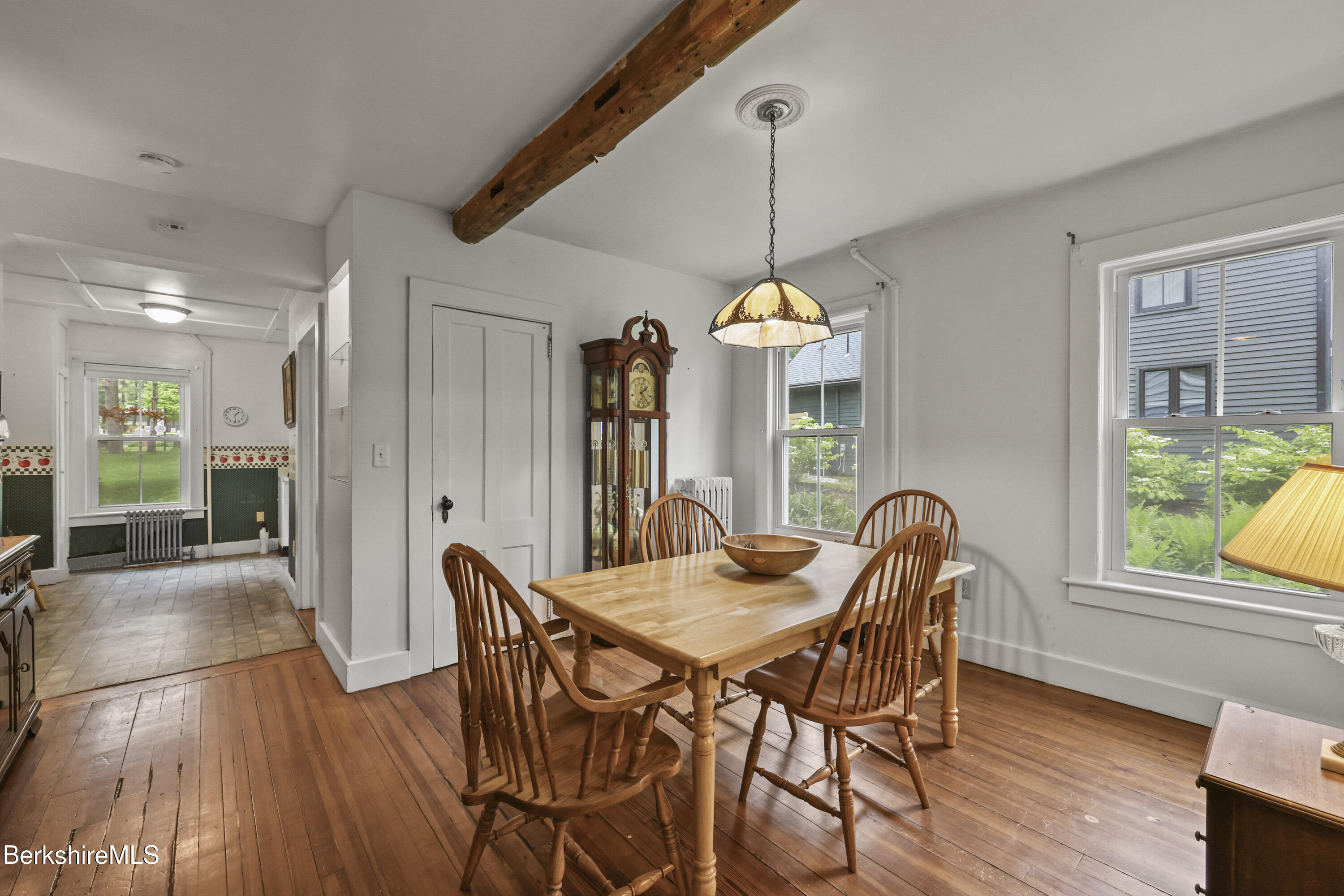 Dining room with a wooden table, exposed beam, grandfather clock, and adjoining tiled kitchen.