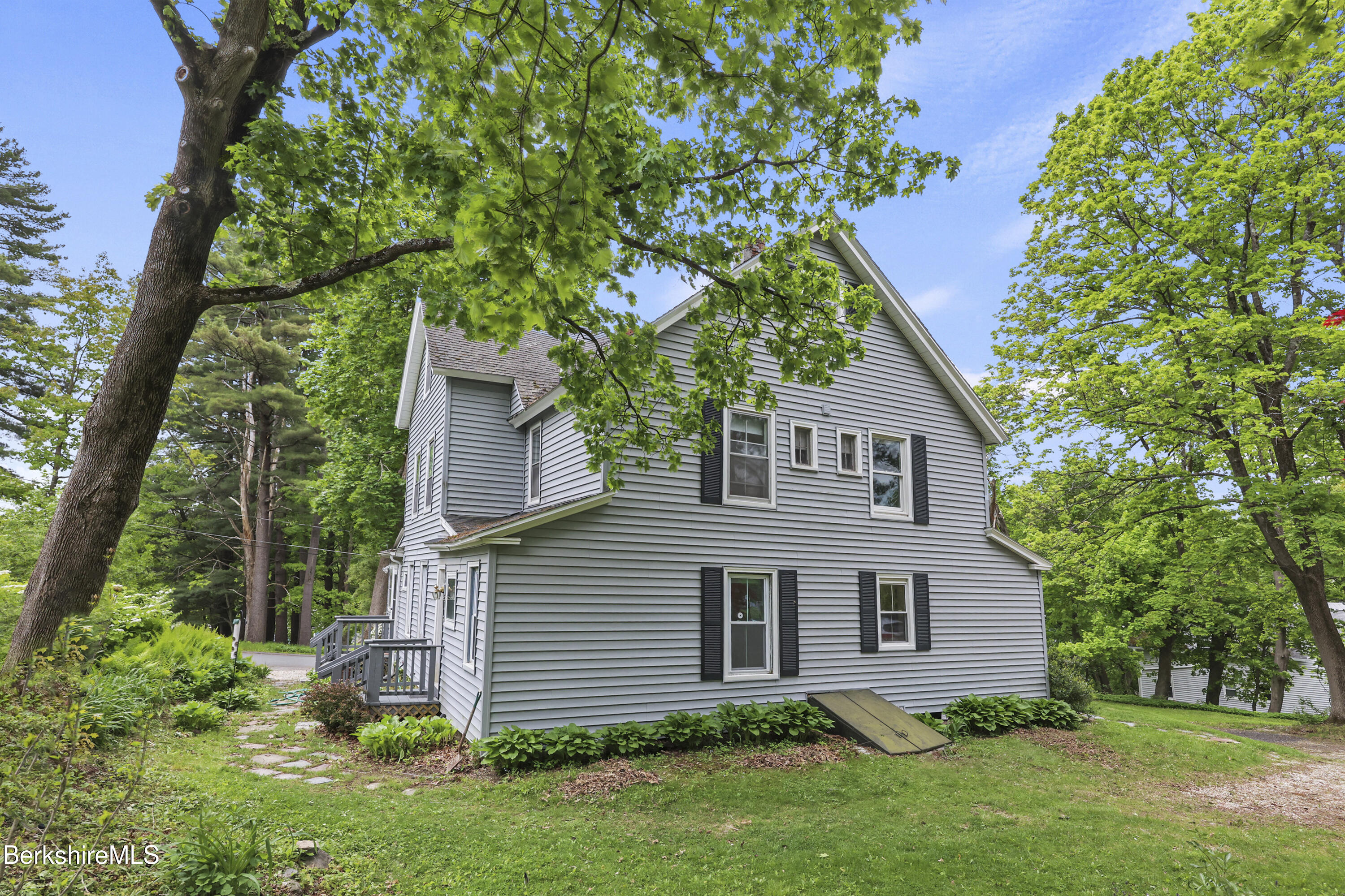 Gray-sided gabled house beneath mature trees, with a small porch and leafy garden beds.