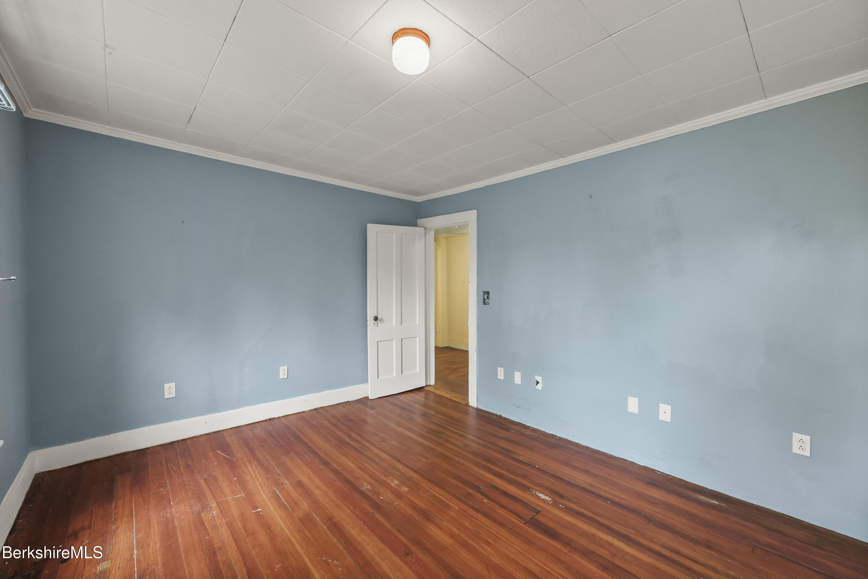 Empty blue-walled room with worn hardwood floors and an open door to a yellow hallway.