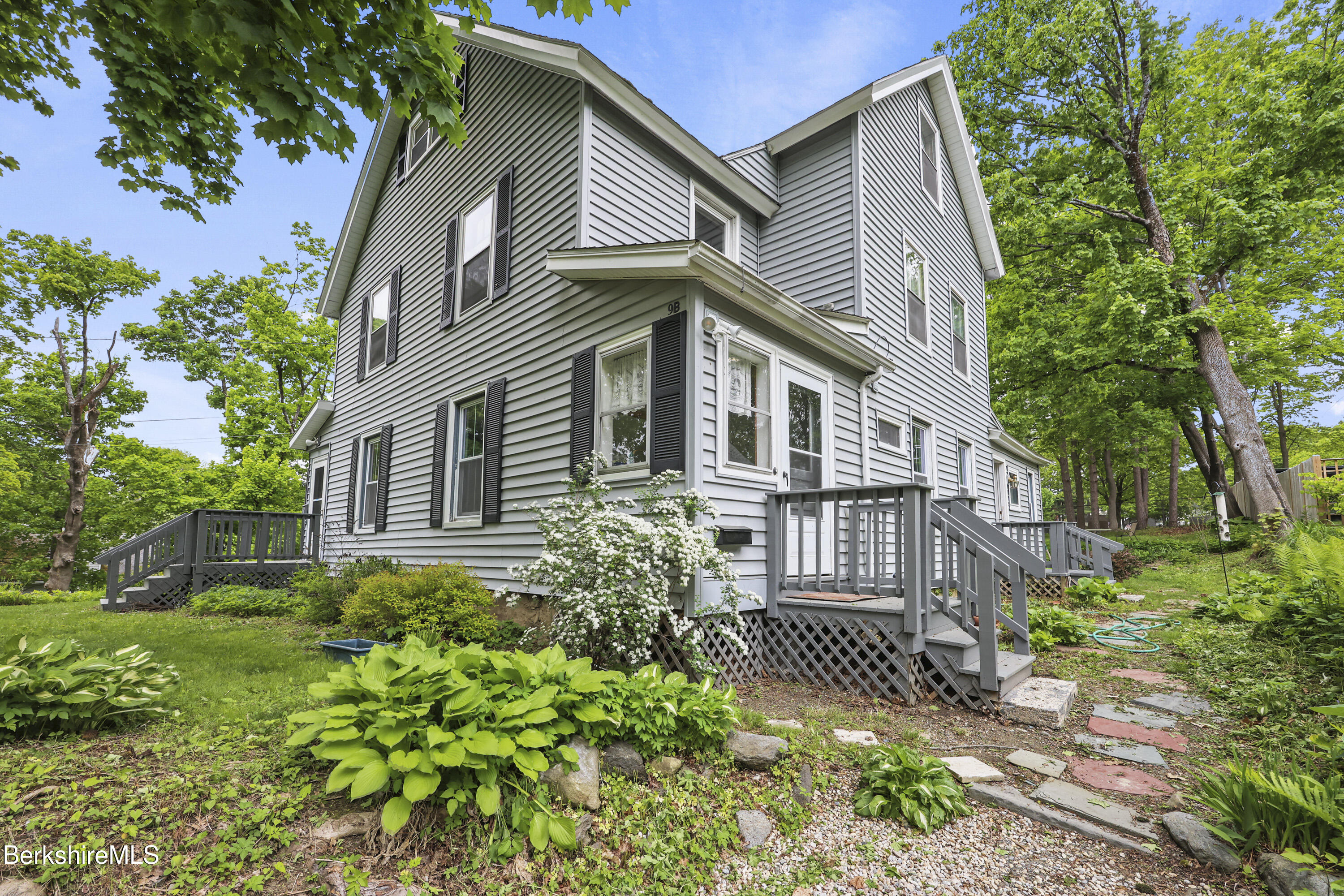 Gray-sided house with black shutters, gray porch steps, and a stone path through a leafy garden.