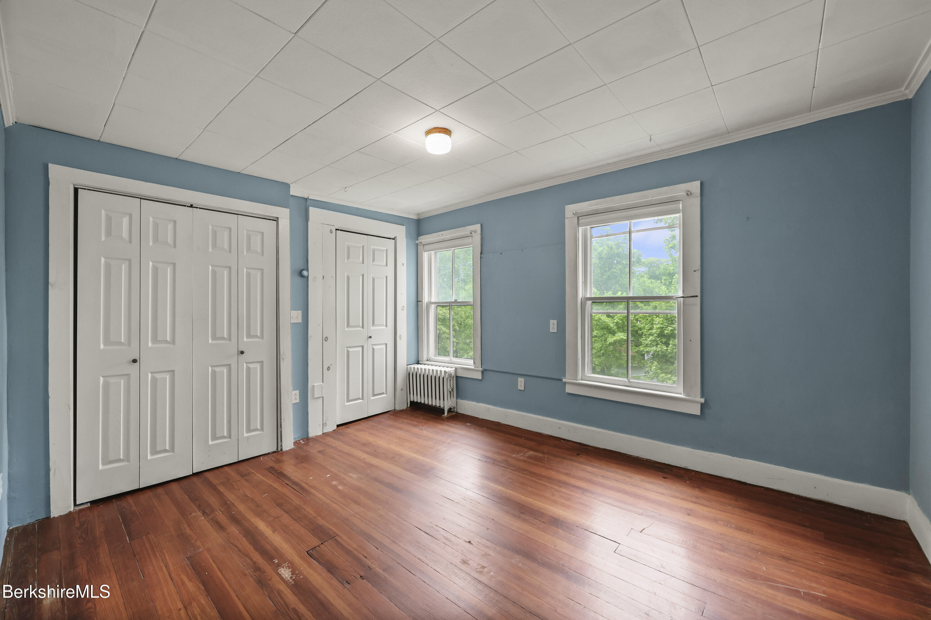 Empty blue room with hardwood floors, white closet doors, and two windows overlooking trees.