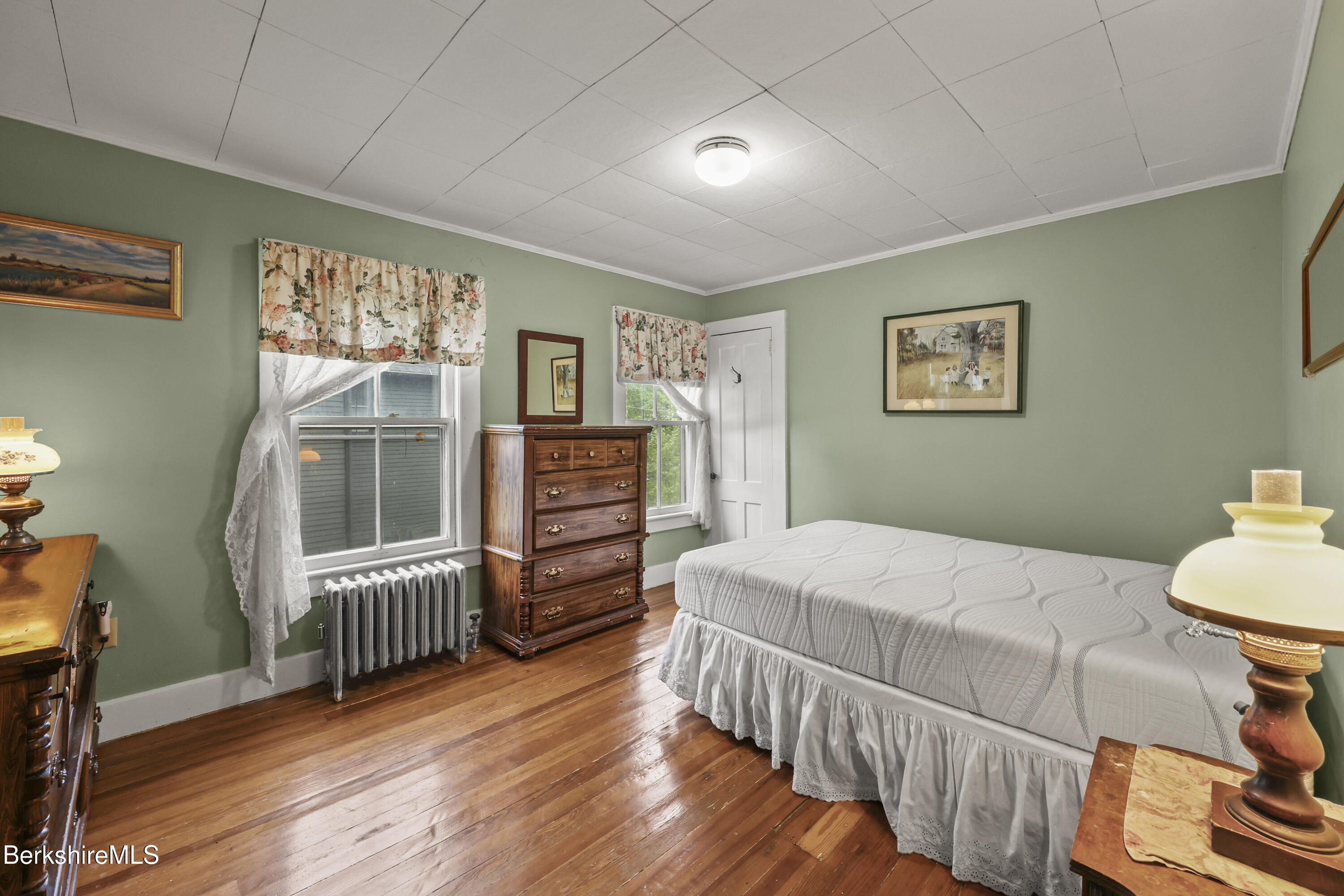 Bedroom with sage-green walls, polished hardwood floors, floral curtains, and a tall wooden dresser.