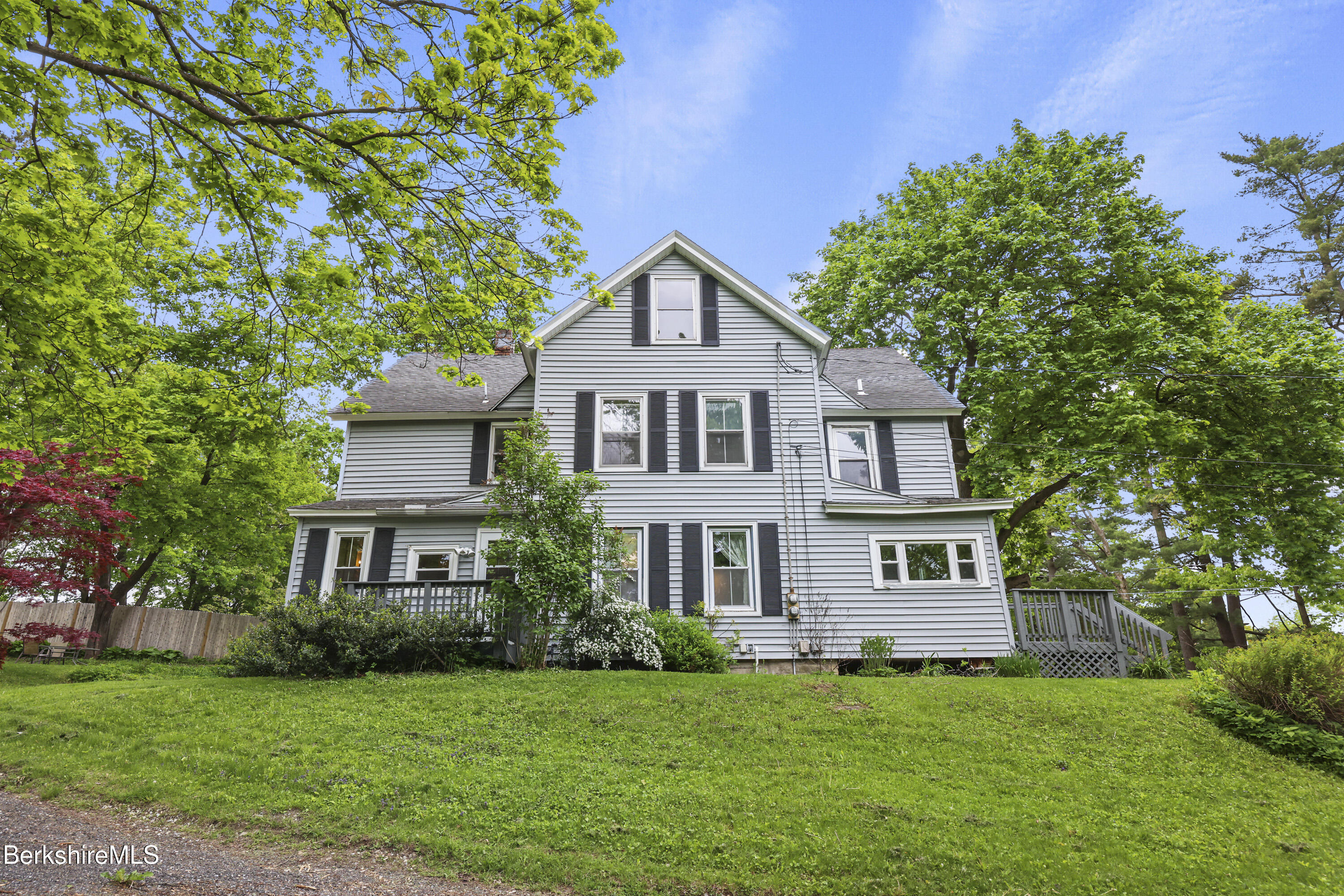 Gray two-story house with black shutters, a porch, and a deck among leafy trees.